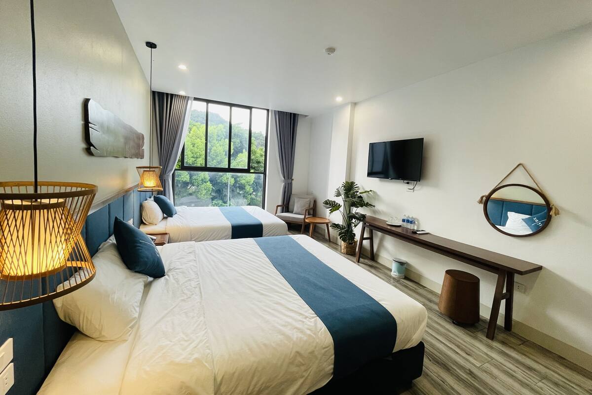 Habitación de hotel moderna con dos camas, un escritorio, televisor y un ventanal que ofrece vistas a zonas verdes.