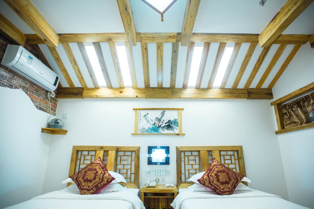 A hotel room with two beds, wooden beams on the ceiling, a wall-mounted air conditioner, and a painting above the beds.