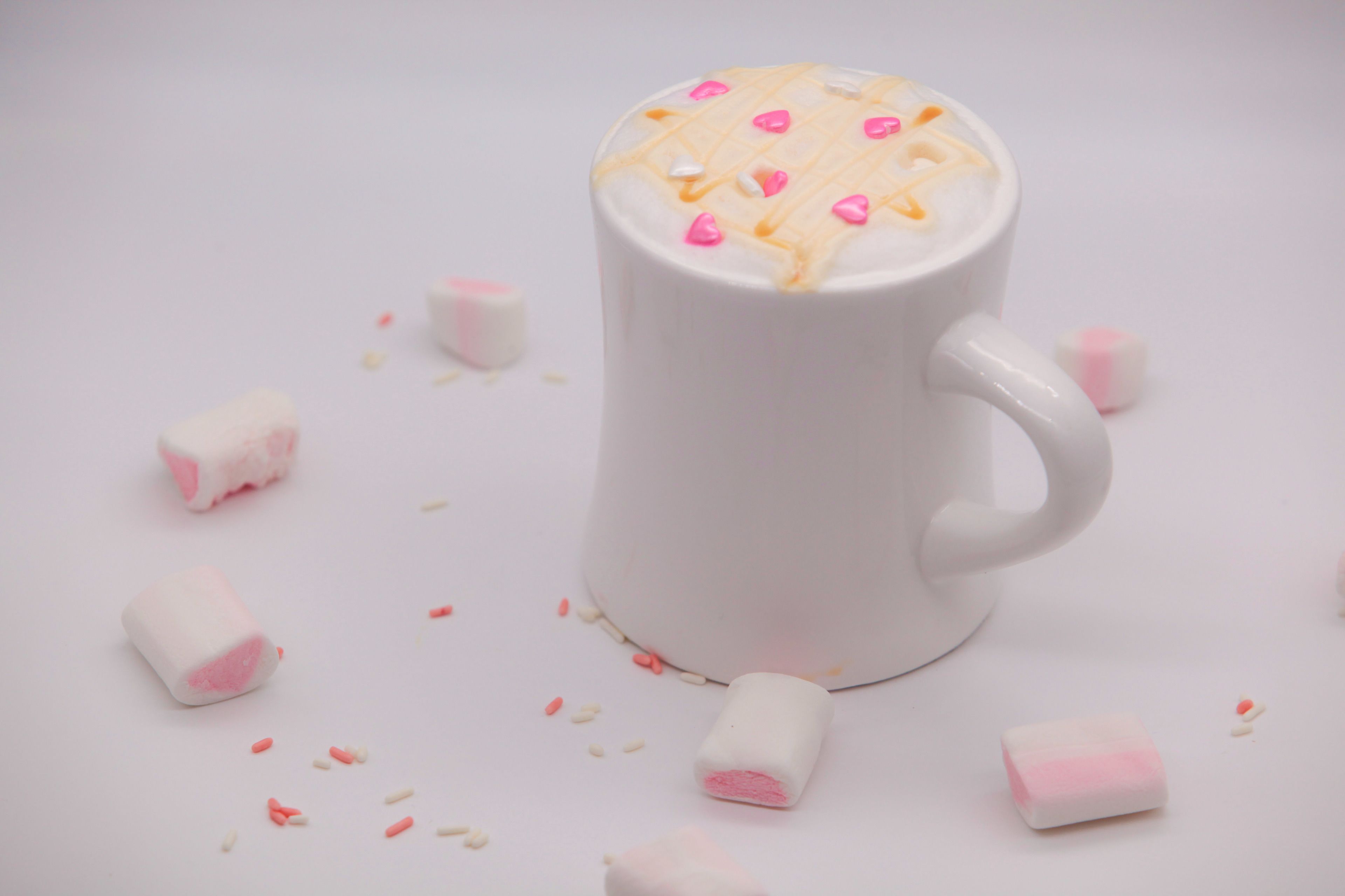 A white mug with a heart-shaped design on the top, surrounded by scattered marshmallows.