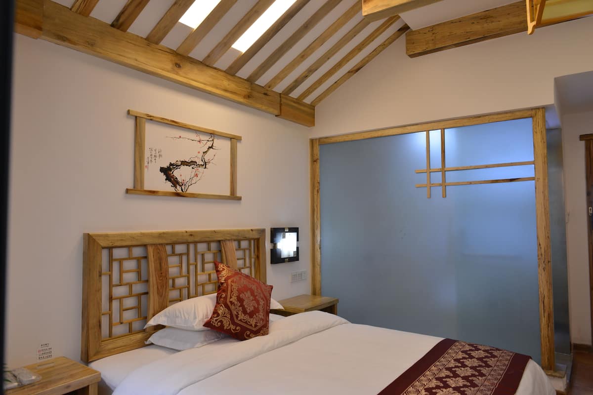 A bedroom with a wooden headboard, a bed with white bedding and a red patterned pillow, a nightstand with a phone, and a framed artwork on the wall.