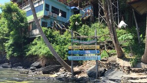 Un cartel con múltiples indicaciones direccionales en un entorno natural, con un río y edificios al fondo.