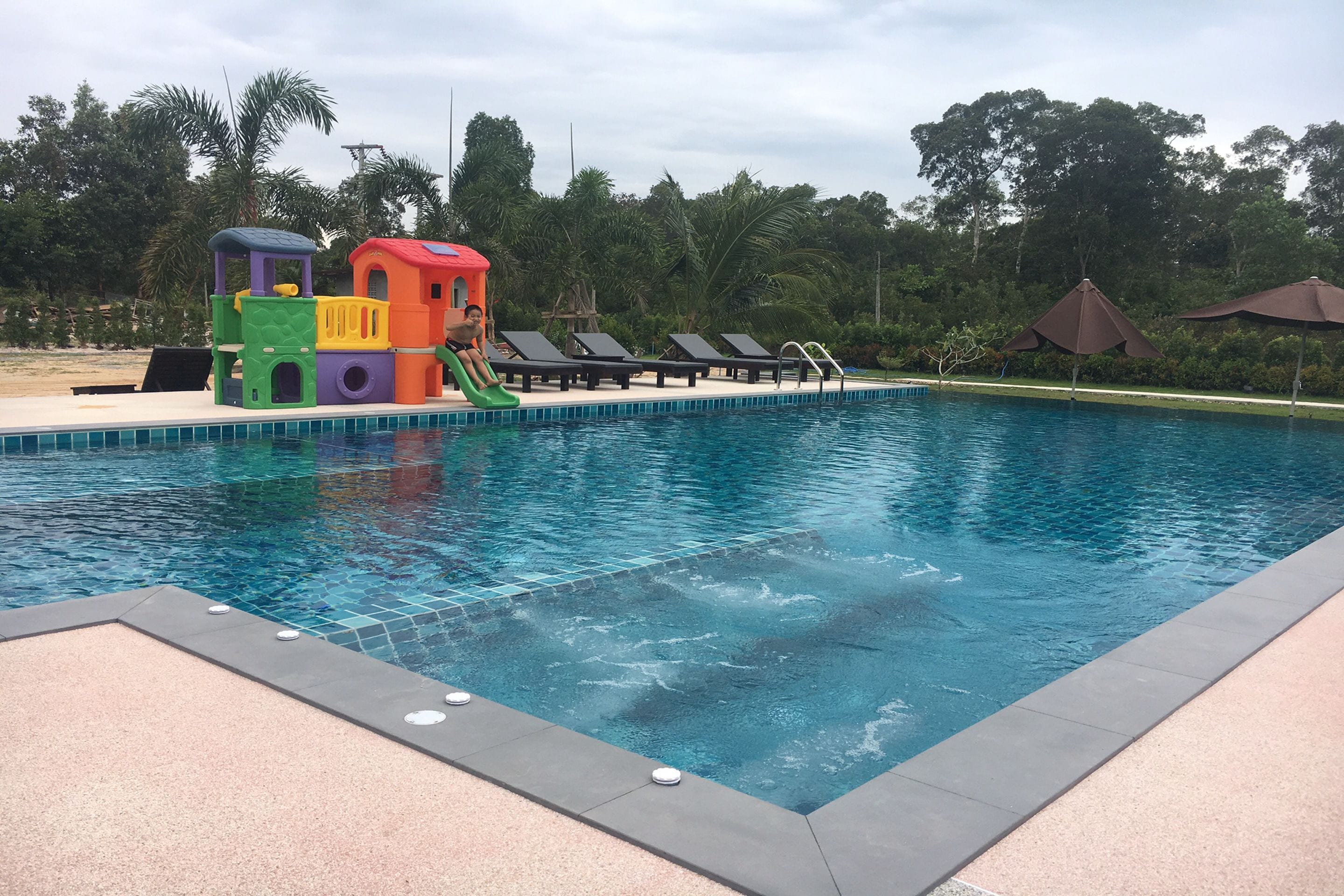 A swimming pool with a slide and play structures, surrounded by lounge chairs and umbrellas.