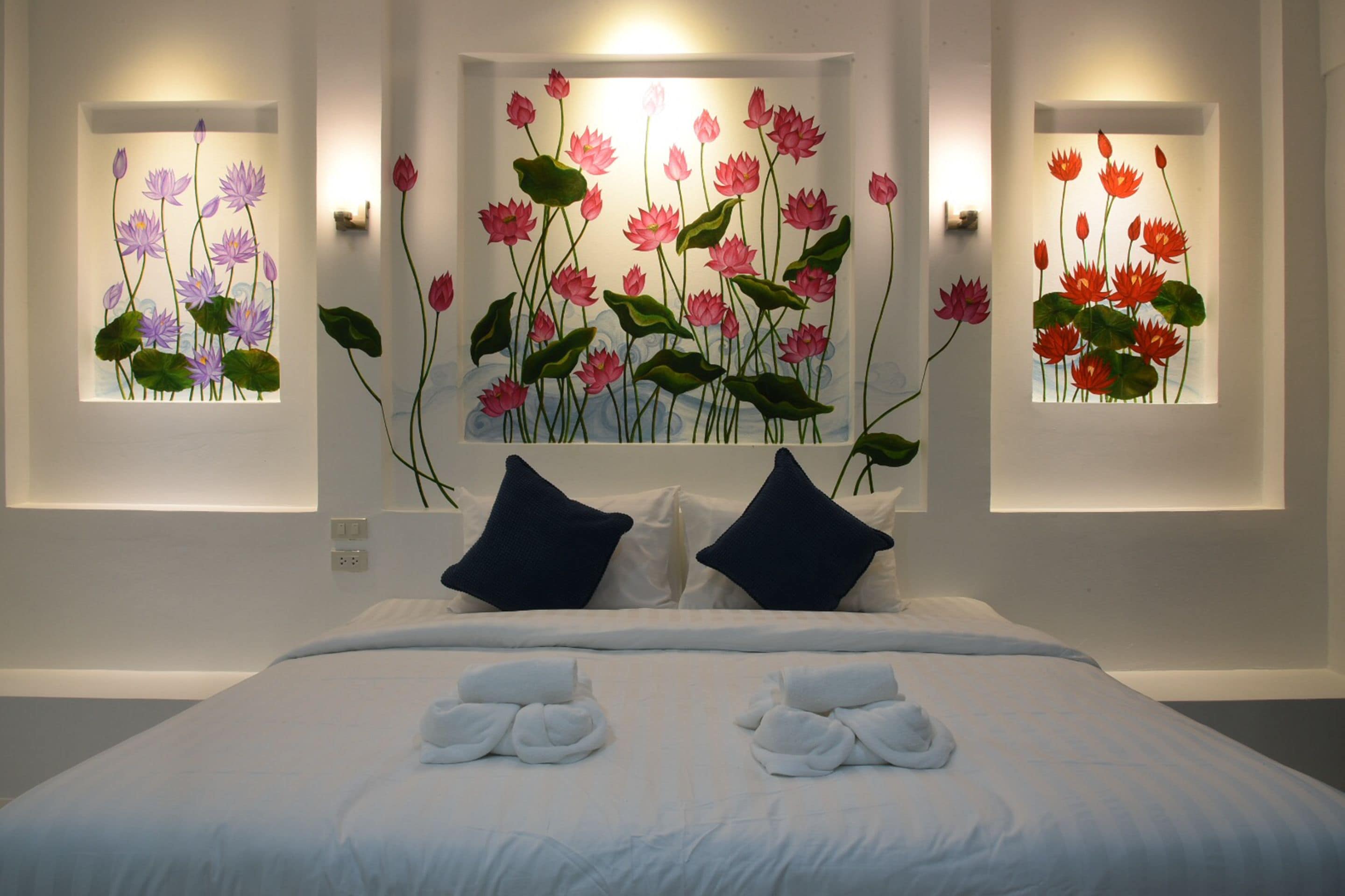 A hotel room with a large bed, two dark blue pillows, and a wall-mounted artwork of pink lotus flowers.
