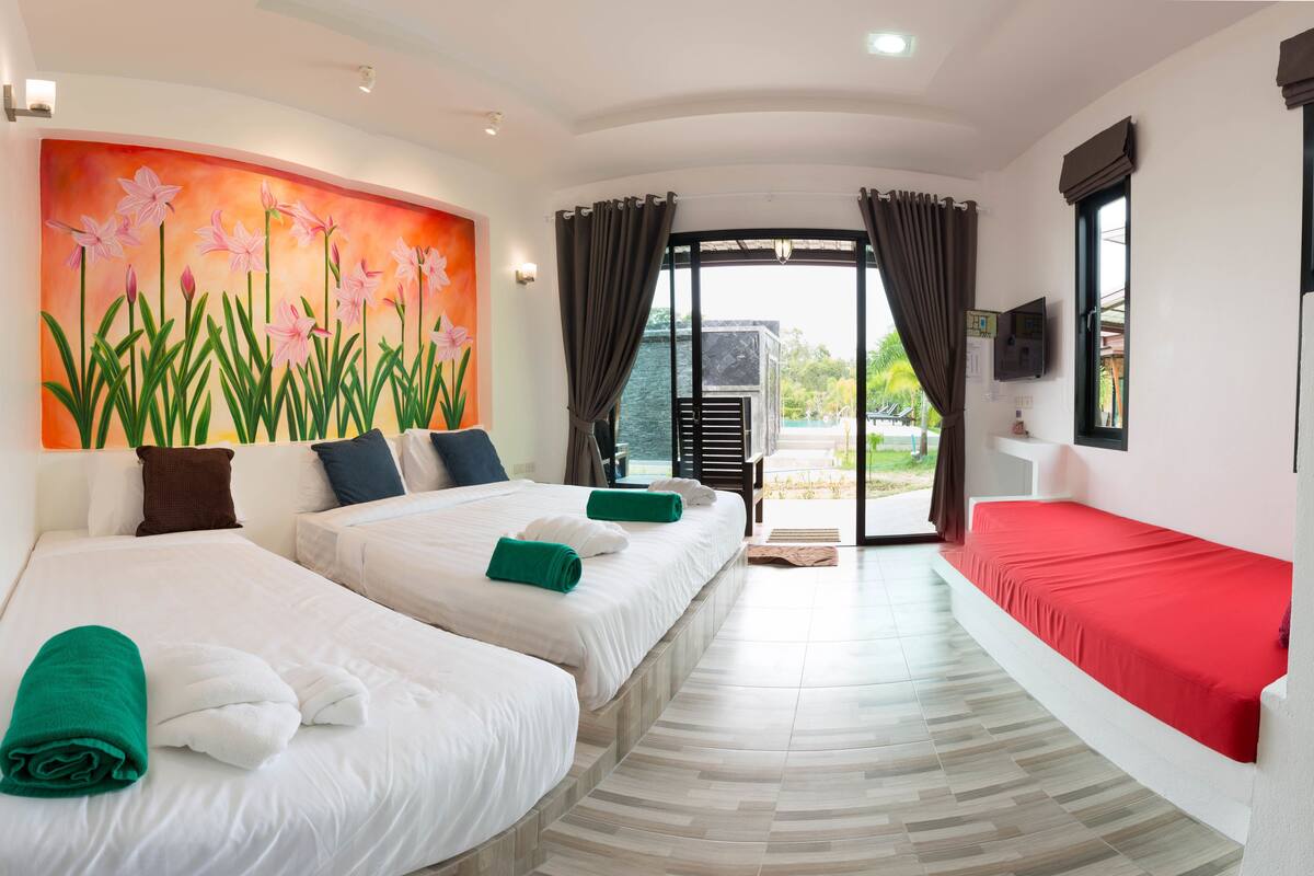 A hotel room with two beds, a painting of flowers on the wall, a view of the outdoors, and a red bedspread.