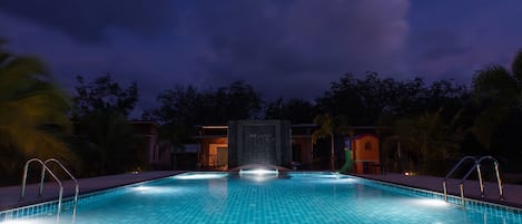 Una piscina con superficie de azulejos, rodeada de palmeras y un edificio al fondo.