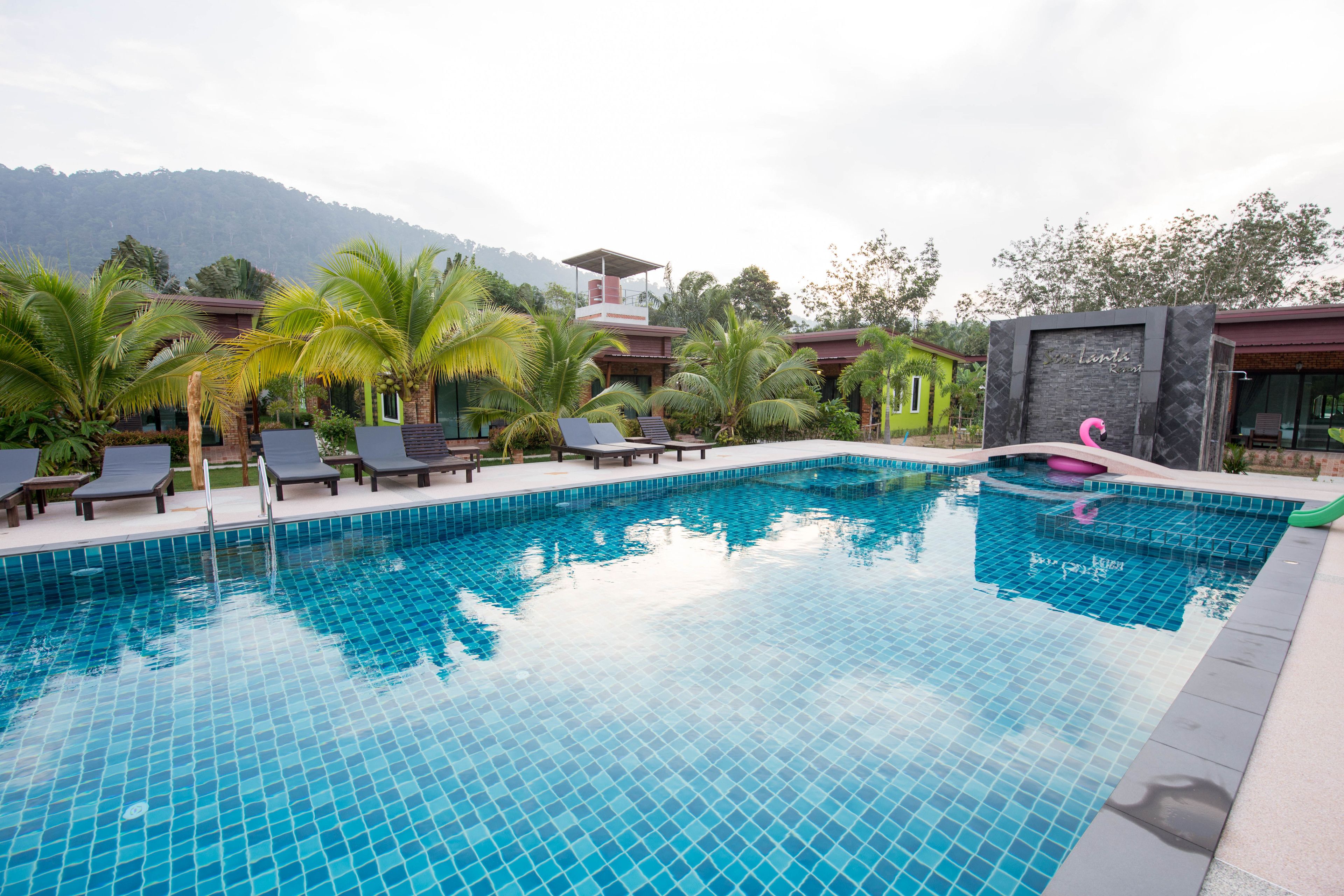 Una piscina con tumbonas, palmeras y montañas al fondo.