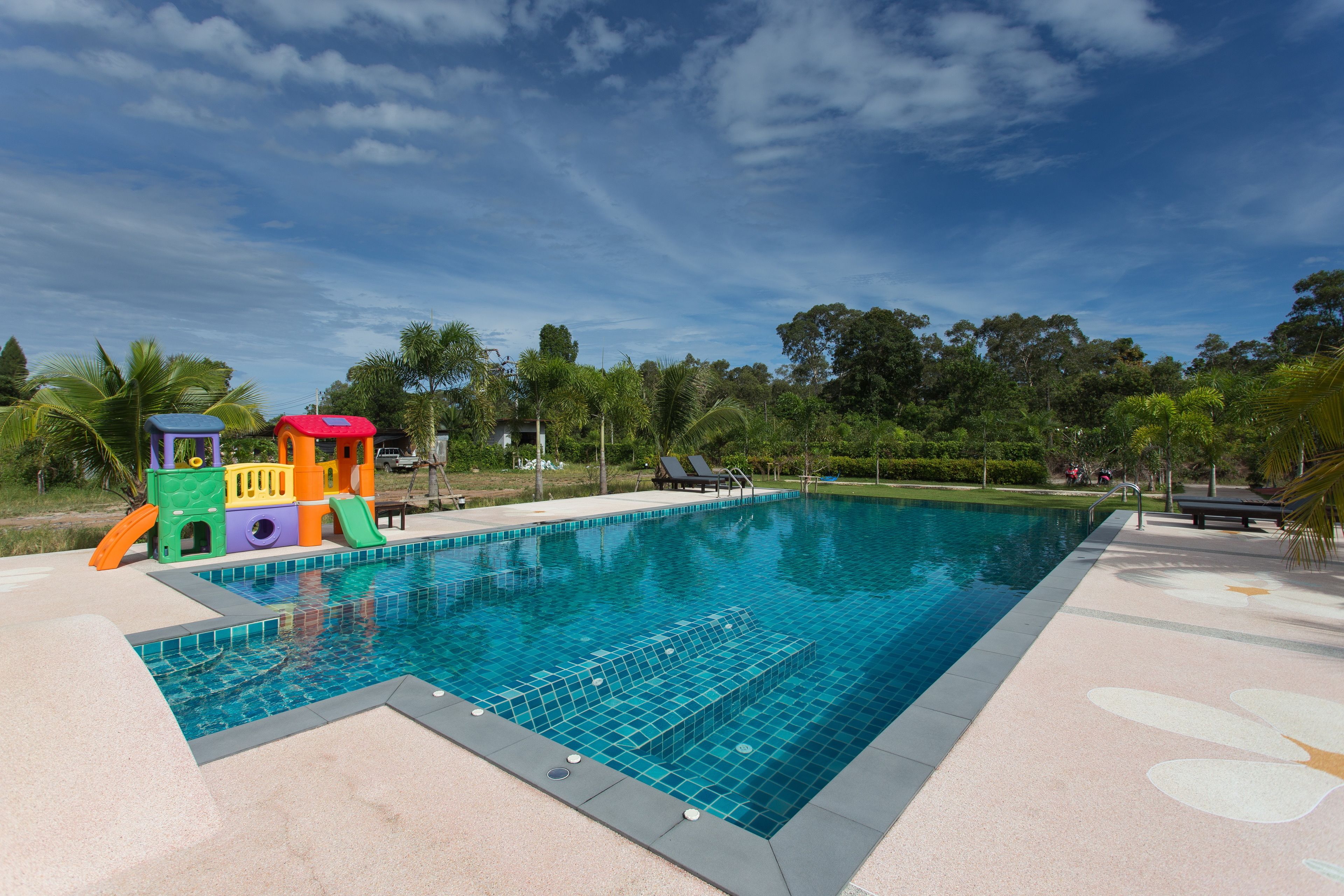 A swimming pool with a children's play structure, surrounded by palm trees and a clear sky.