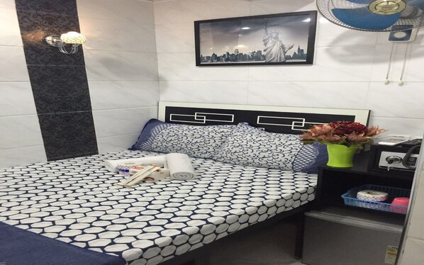A neatly arranged hotel room with a patterned bedspread, a framed cityscape picture, a bedside fan, and a small table with a vase of flowers.