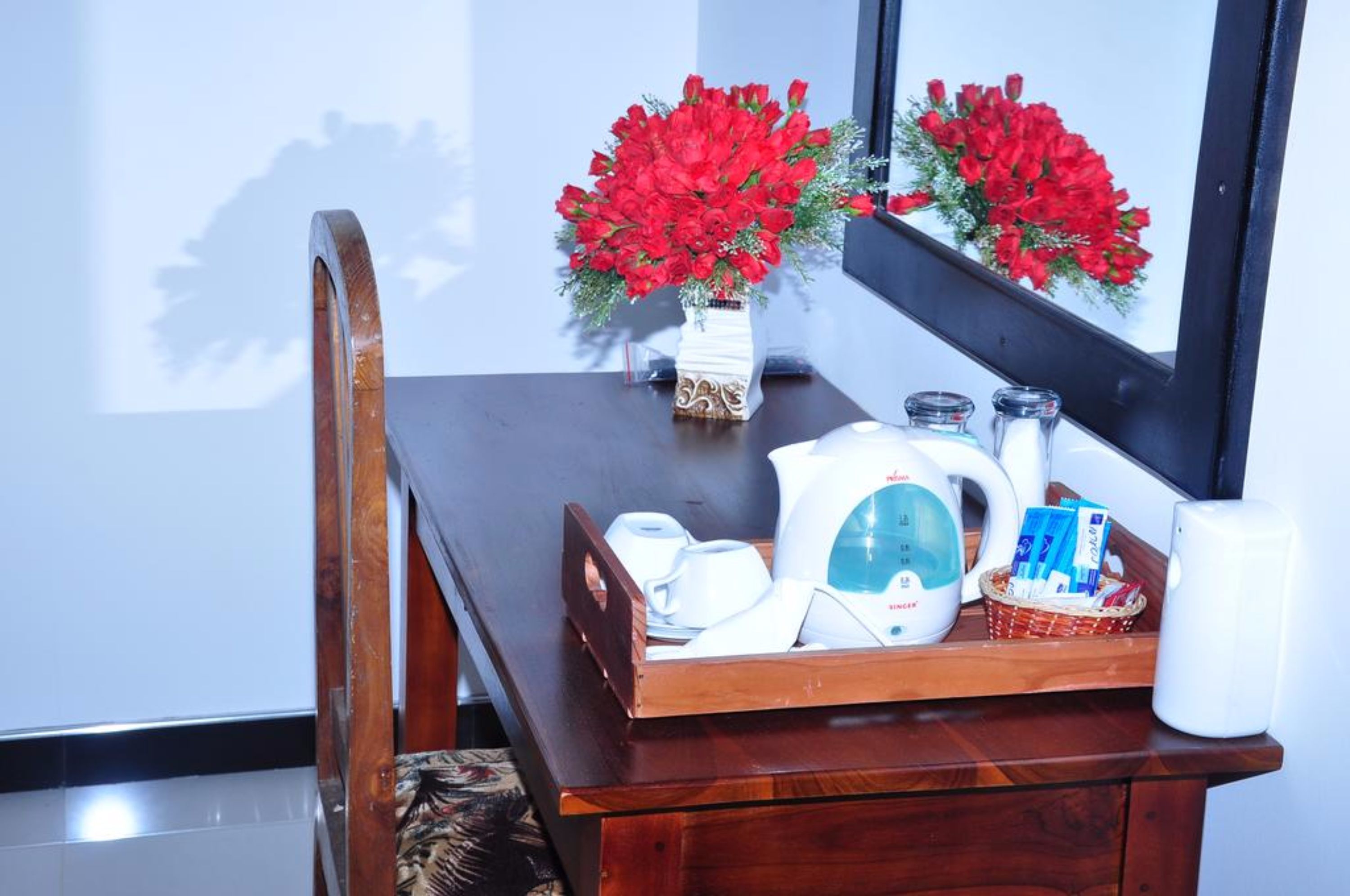 A wooden dining table with a white vase of red flowers, a white kettle, a basket with items, and a mirror reflecting the flowers.