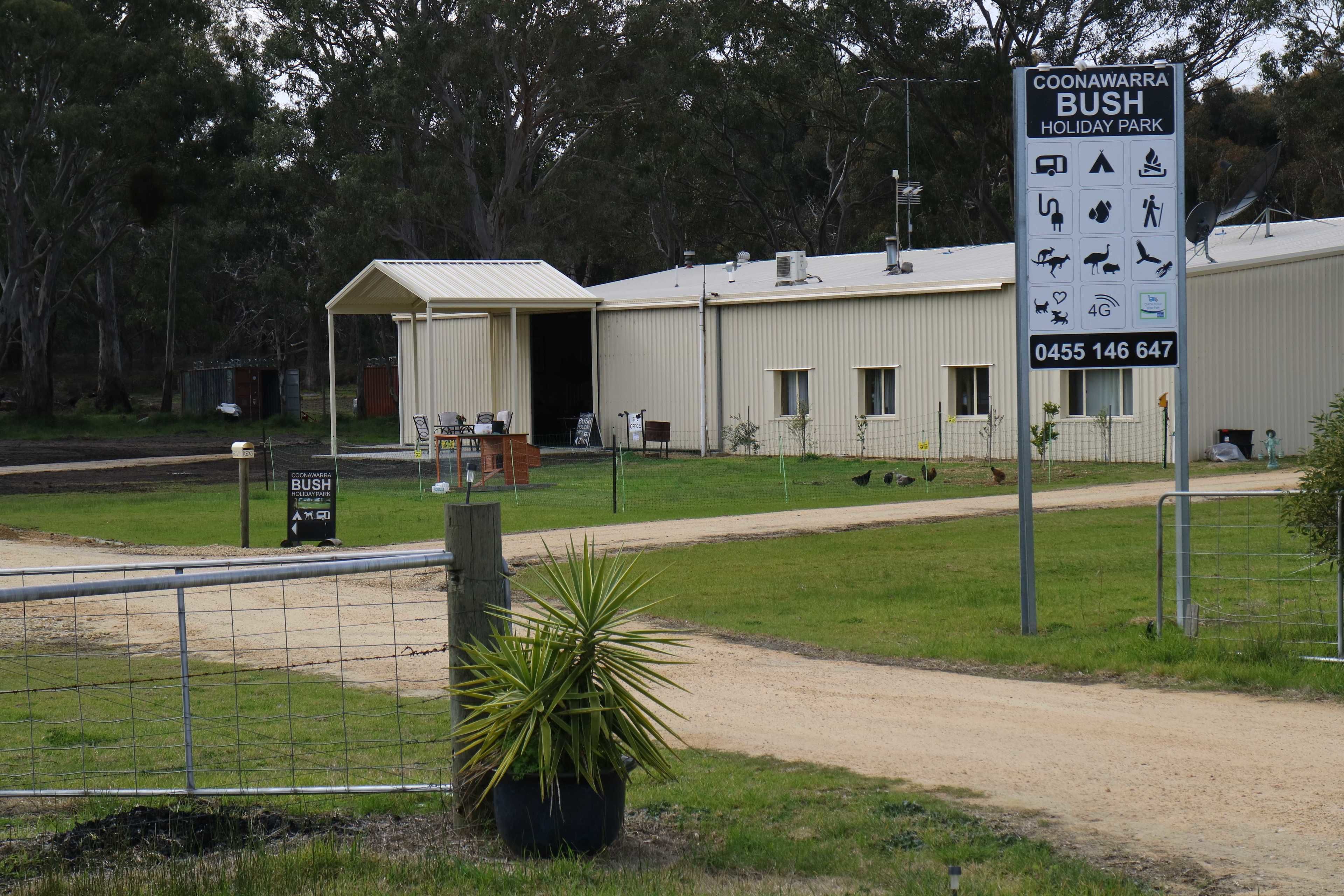 A sign for Coonawarra Bush Holiday Park with amenities icons, a contact number, and a fenced area with a building and a potted plant.