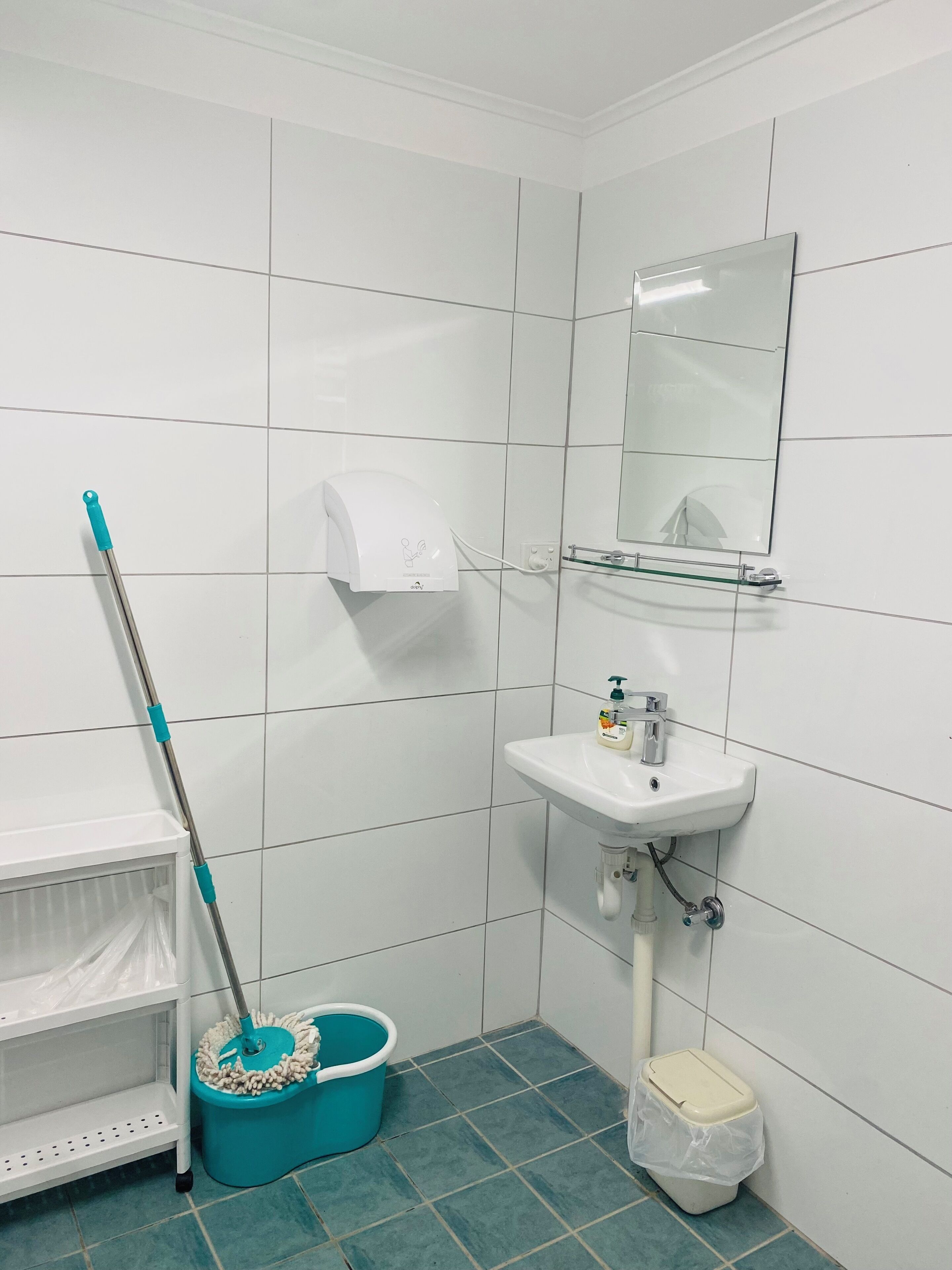 Une salle de bain avec des carreaux blancs, un lavabo, un miroir, une serpillière, un seau et une poubelle.