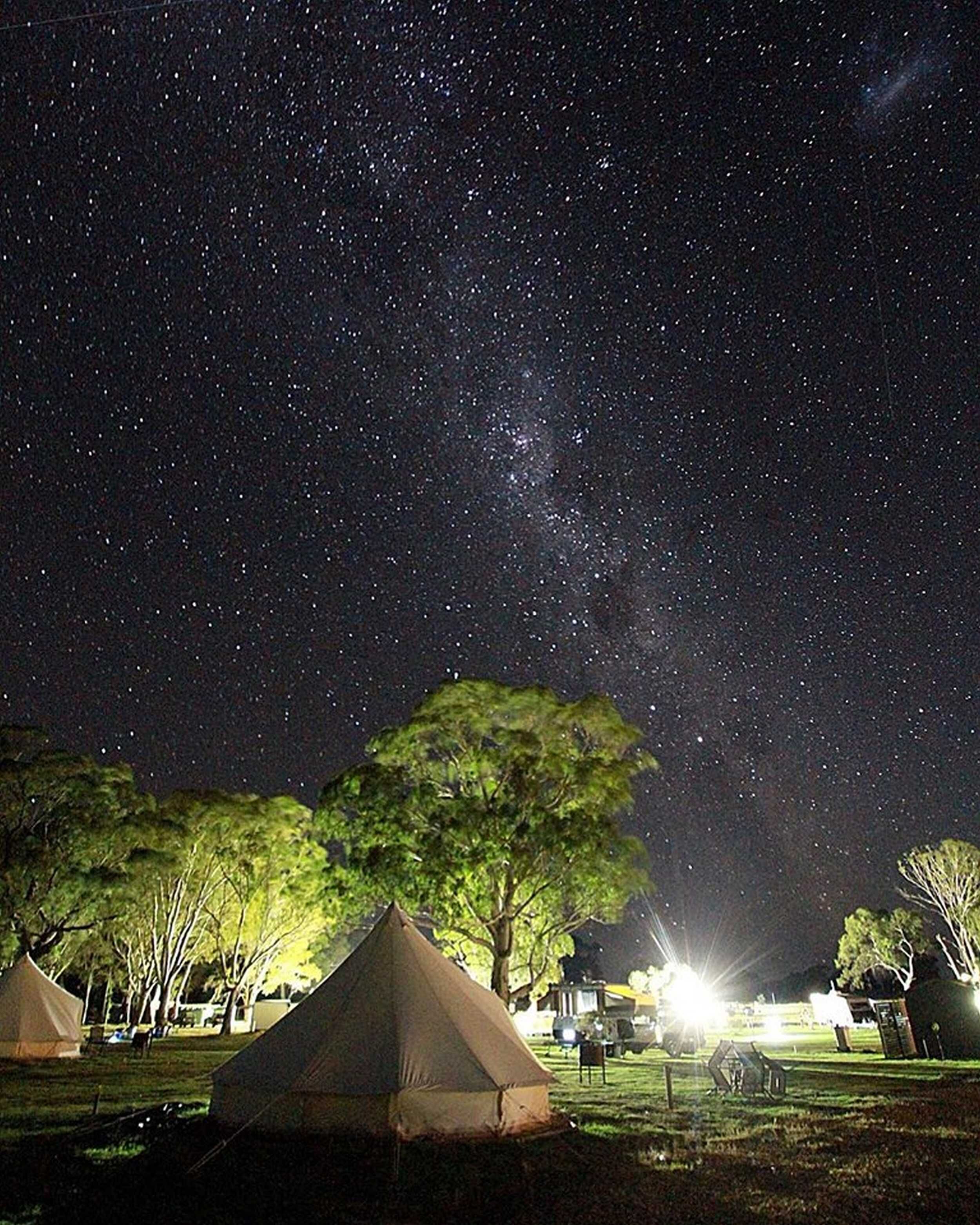 A night sky with visible stars and the Milky Way. There are tents and trees in a grassy area.
