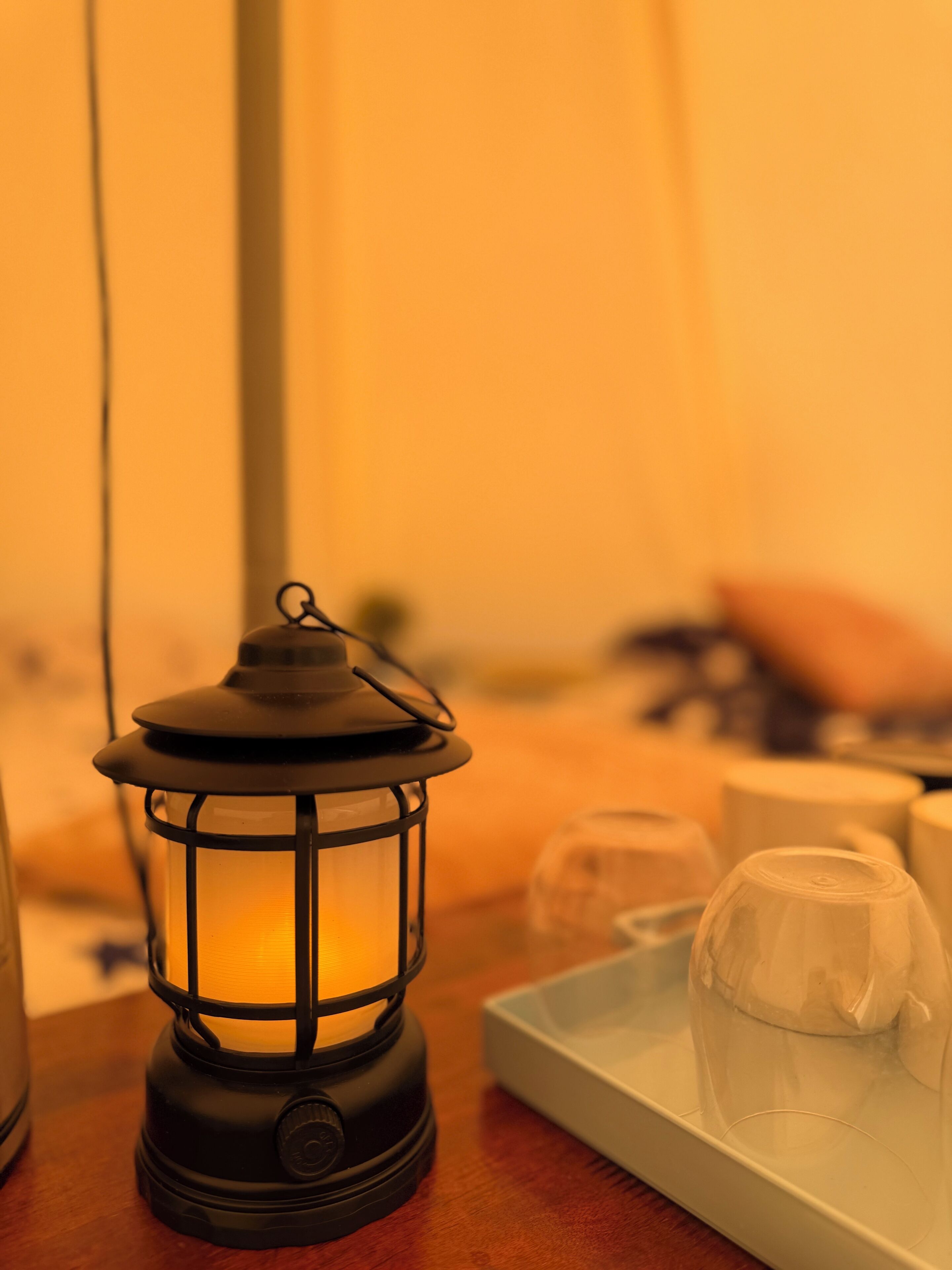 Un farol negro con revestimiento de vidrio, sobre una bandeja que contiene un recipiente de vidrio transparente y un objeto blanco, posiblemente un candelabro, sobre una superficie de madera.