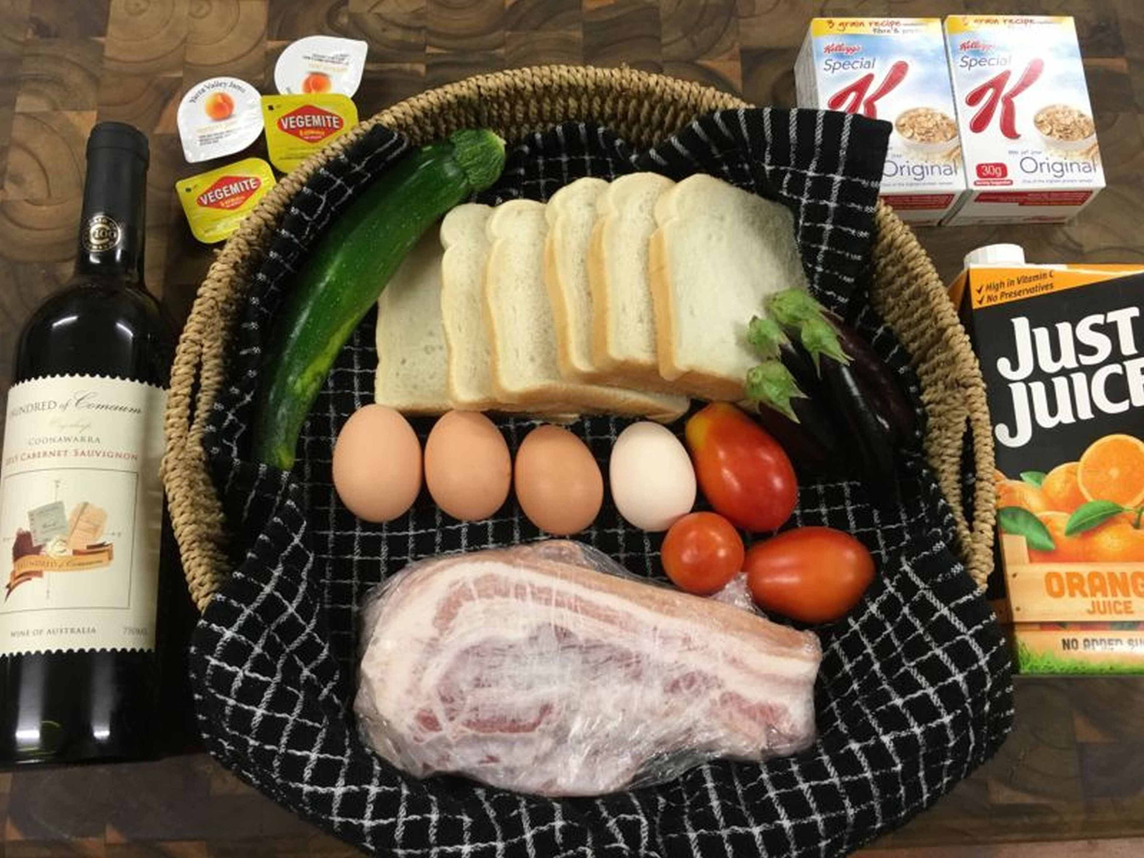 A basket containing bread, eggs, tomatoes, a cucumber, a piece of meat, and a bottle of wine.