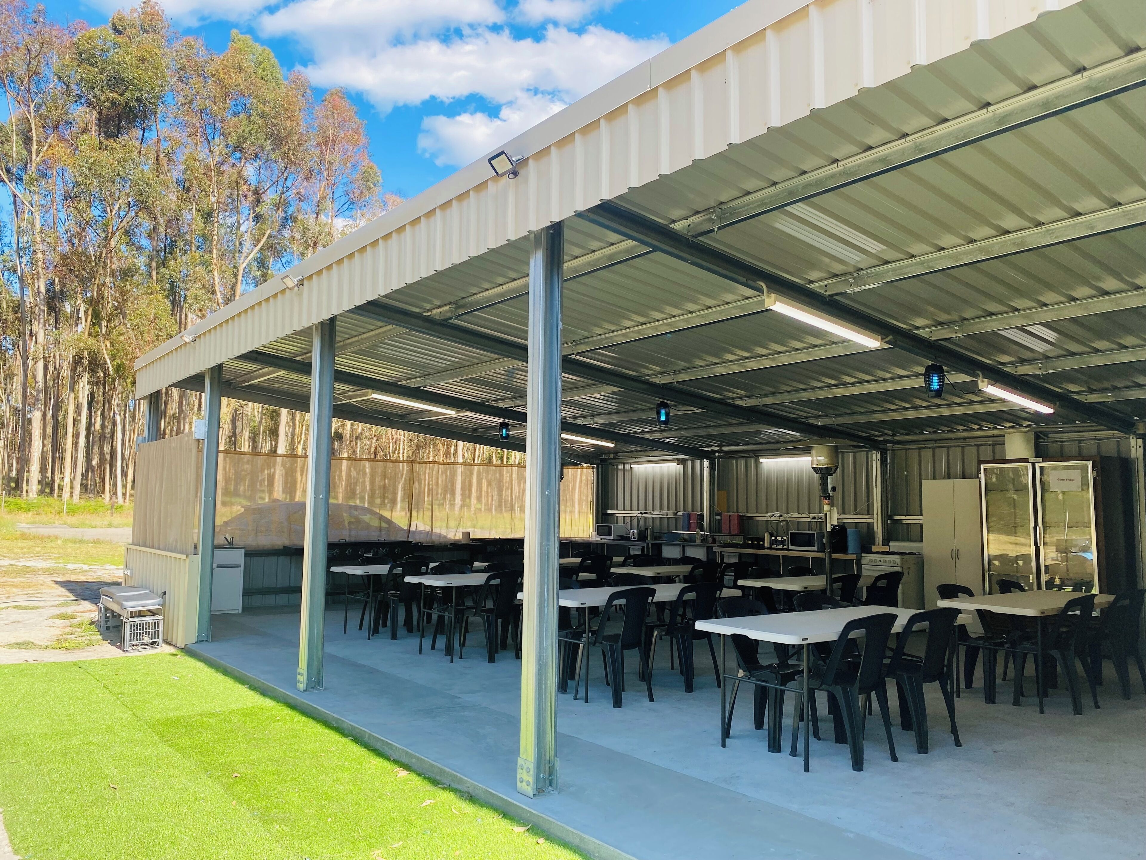 An outdoor covered area with tables and chairs, a large screen, and a fenced area with trees.