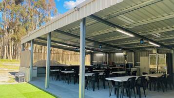 An outdoor covered area with tables and chairs, a large screen, and a fenced area with trees.