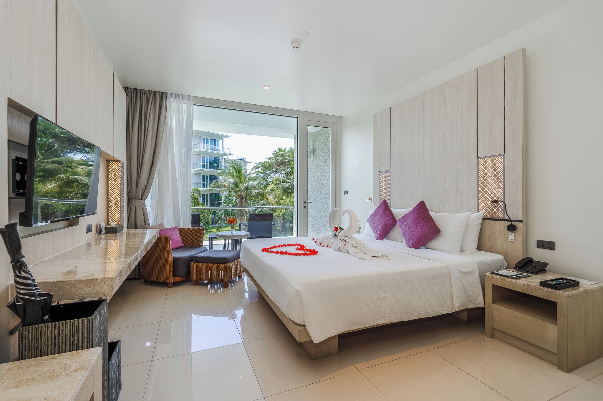 A modern hotel room with a large bed, a TV, a balcony with a view of palm trees, and a dining area with a table and chairs.