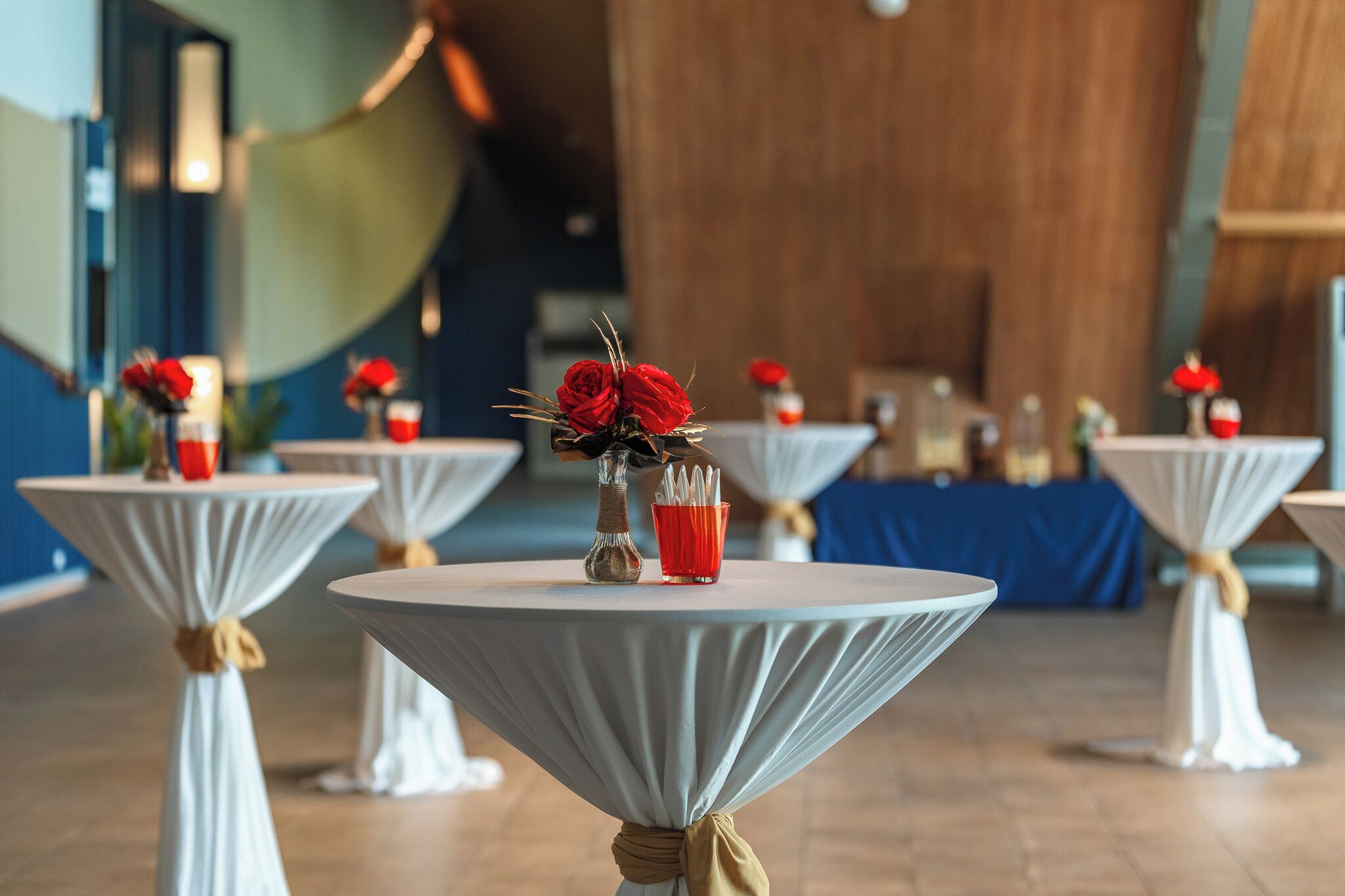 Round tables with white tablecloths and red roses, set in a room with wooden paneling and blue curtains.