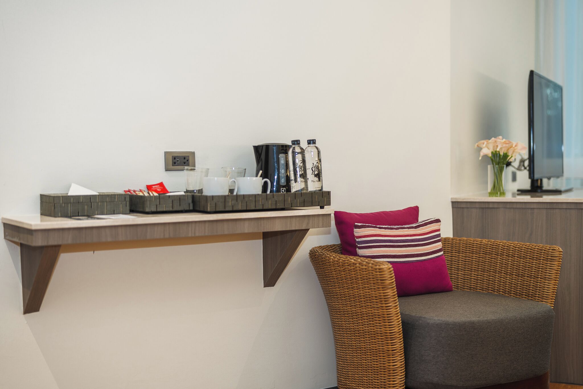 A modern living area with a wicker chair, a coffee station with cups and bottles, and a television on a stand.
