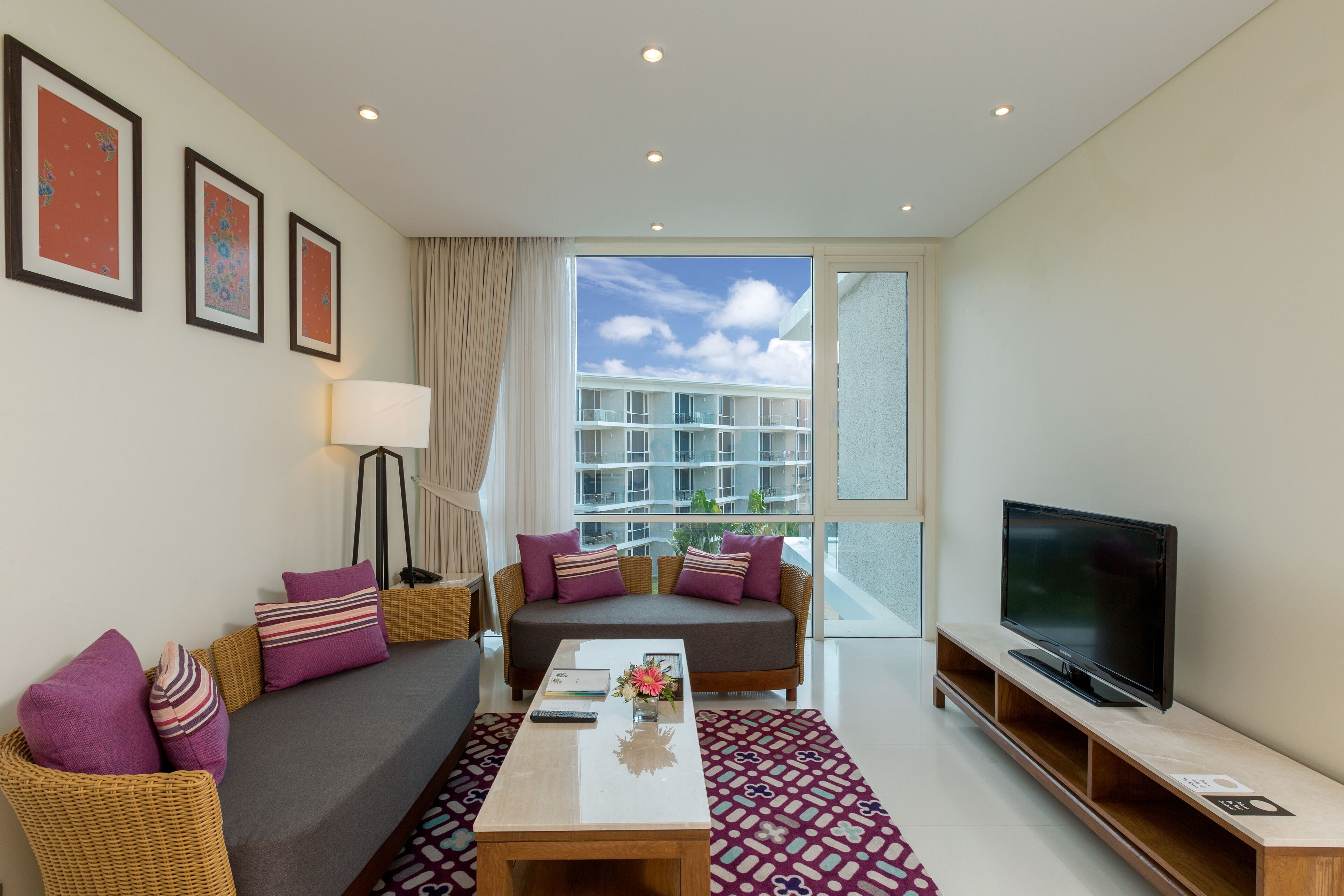 A modern living room with a sofa, coffee table, and TV. There are framed pictures on the wall and a large window with a view of buildings.