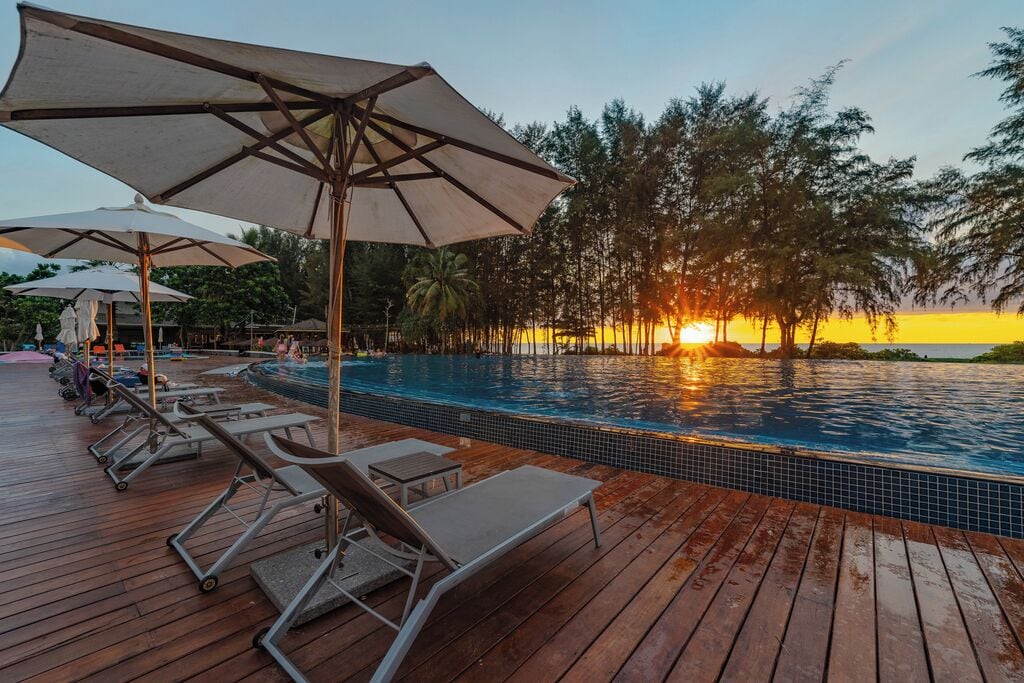 A wooden deck with lounge chairs and umbrellas overlooking a swimming pool at sunset.