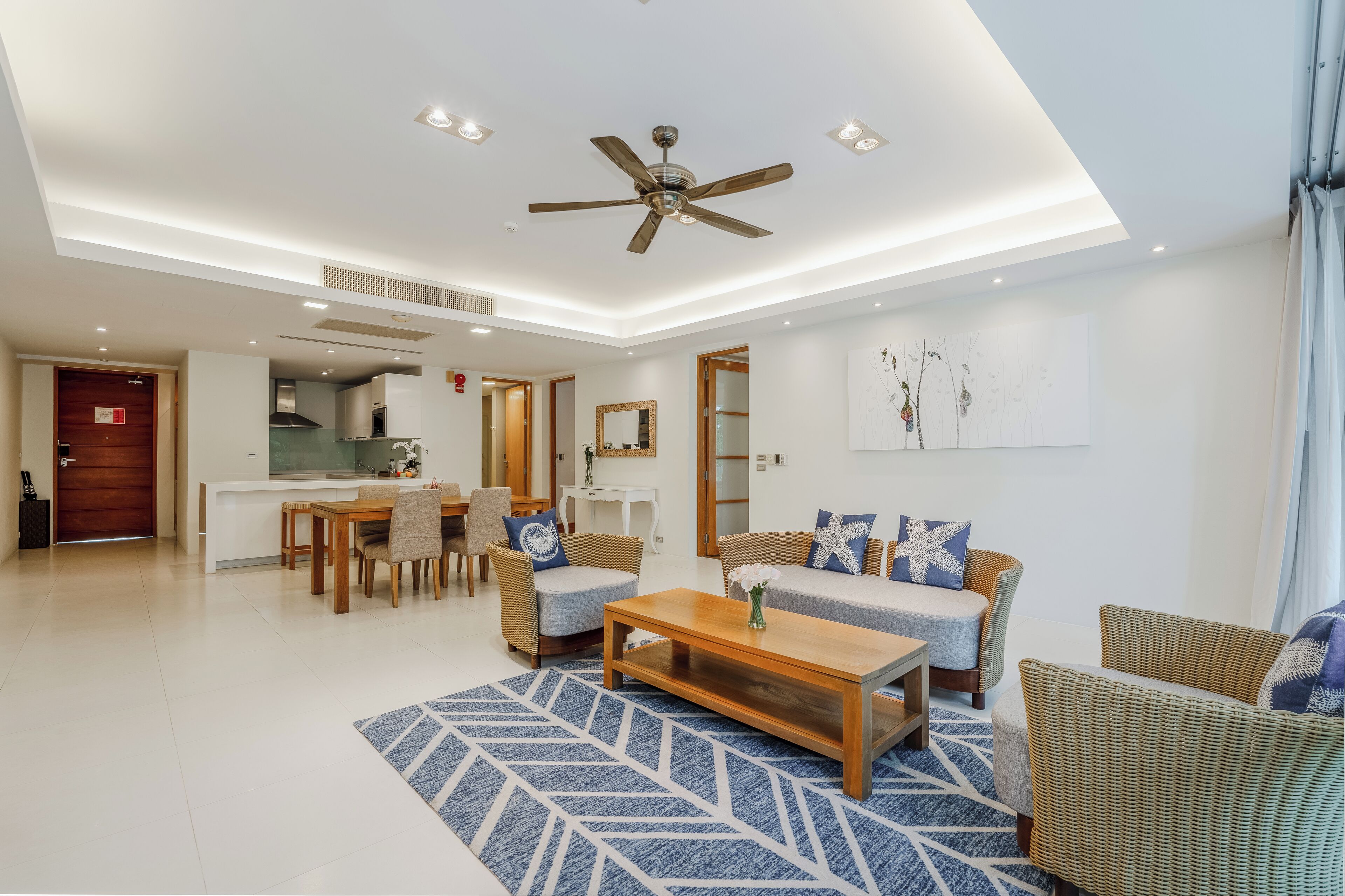 A modern living room with a ceiling fan, a dining area with a table and chairs, a sofa with cushions, and a kitchen in the background.