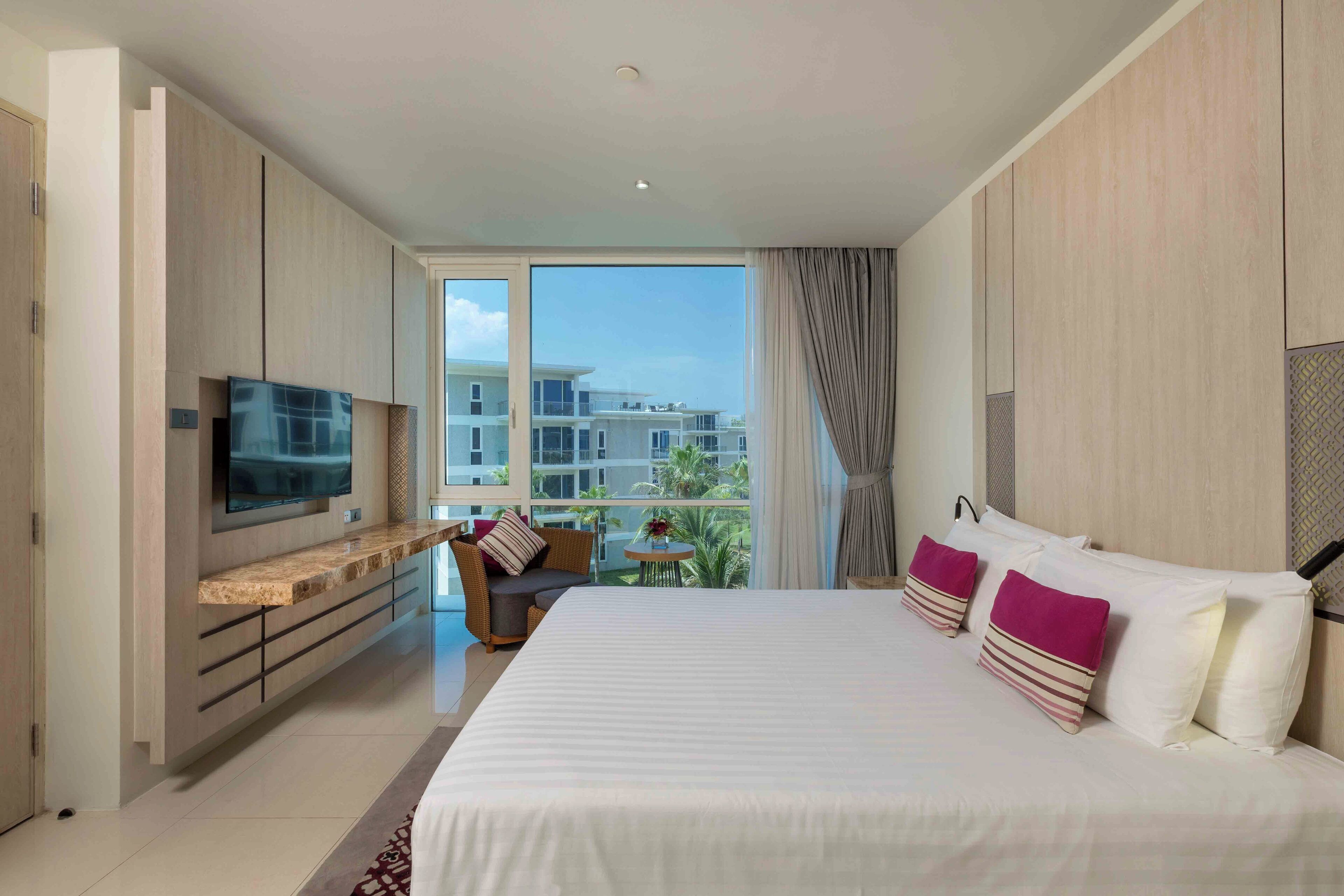 A modern hotel room with a large bed, a TV mounted on the wall, a balcony with a view of buildings, and a small table with a chair.