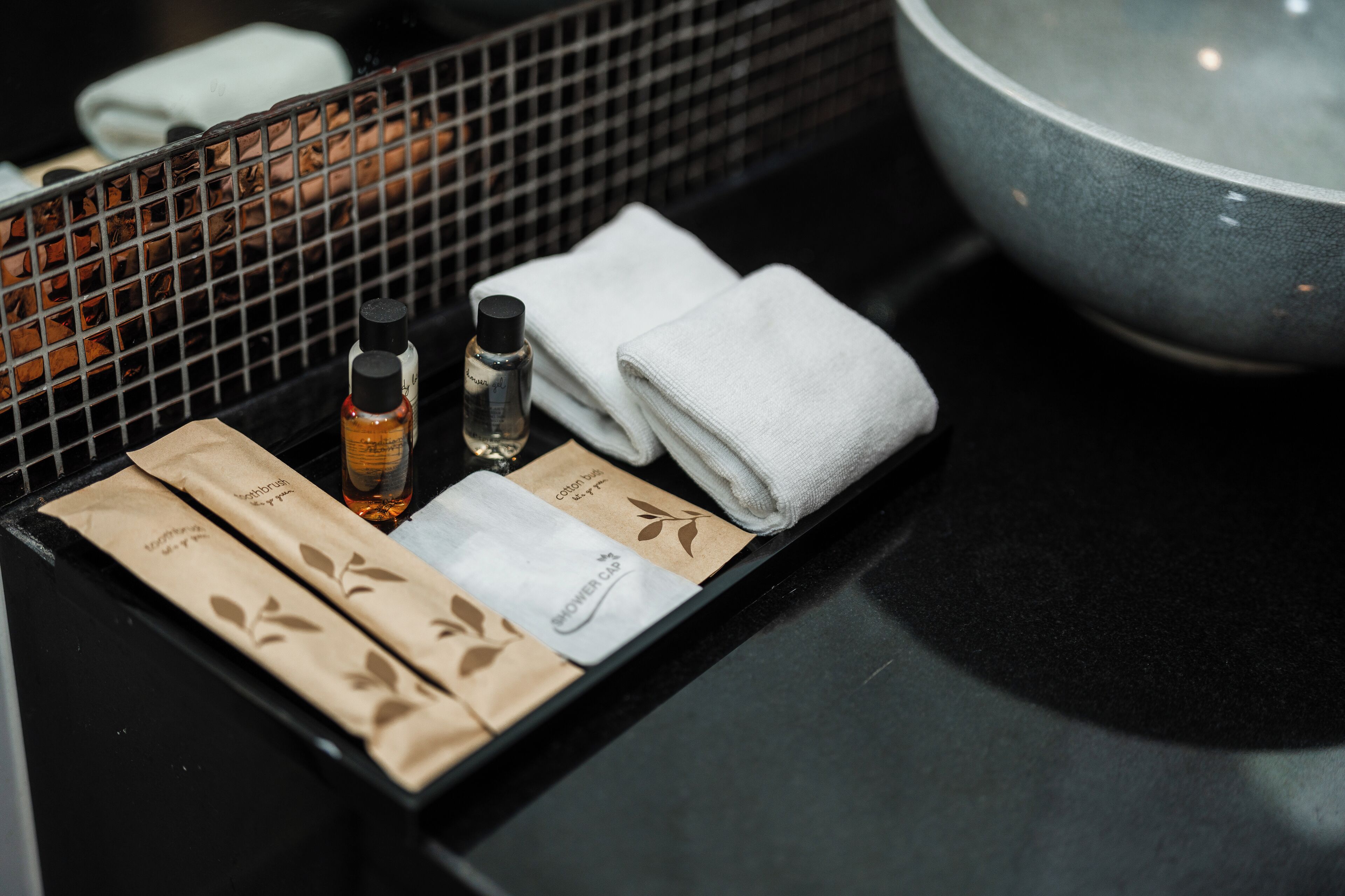 A bathroom counter with a black sink, a large bowl, and toiletries including a bottle, towels, and paper bags.