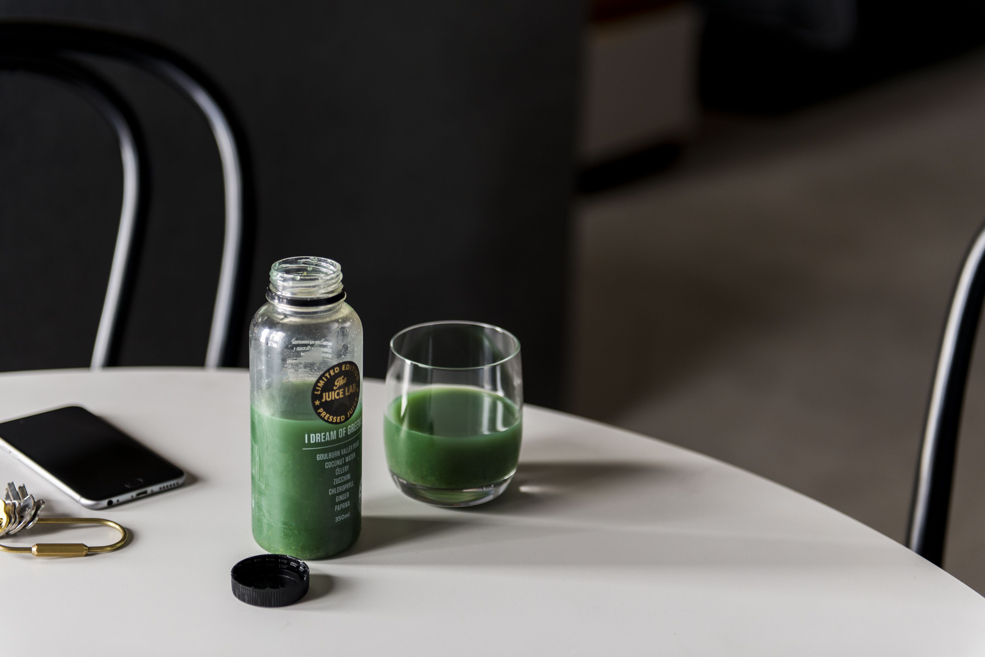 Un vaso de jugo verde, un vaso y un teléfono inteligente sobre una mesa blanca.