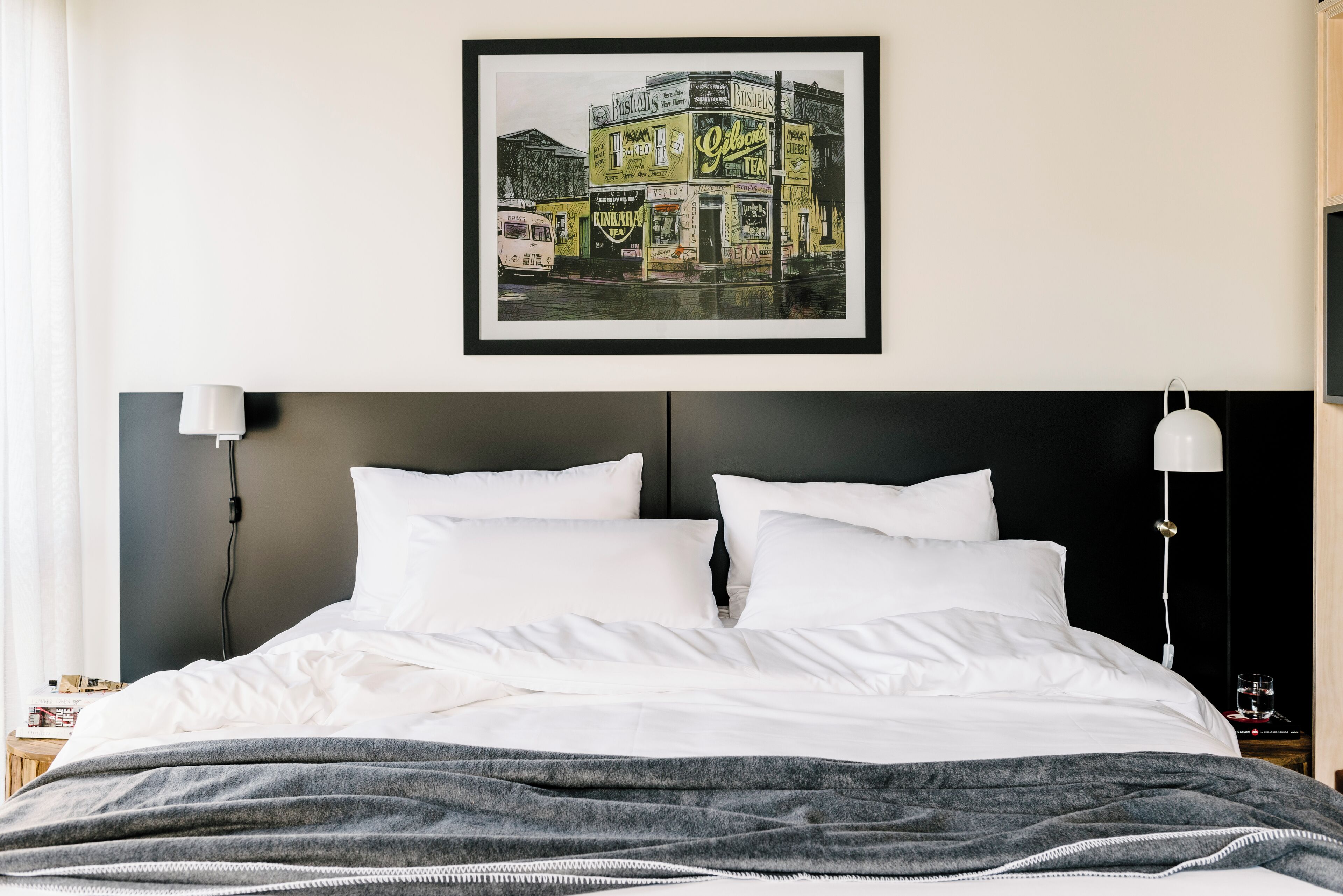 A neatly made bed with white linens and a gray throw blanket, a framed picture of a building on the wall, and two bedside lamps.