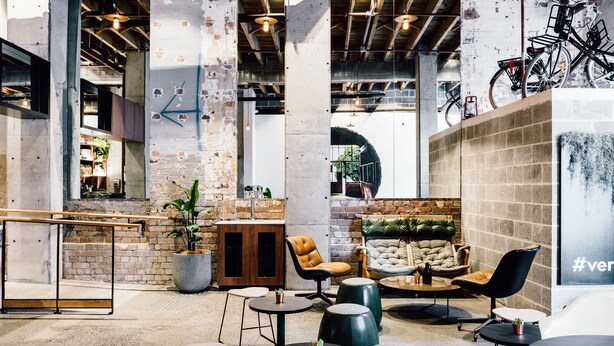 A modern interior with exposed brick walls, concrete pillars, and a mix of seating areas including stools and a sofa. - Veriu Broadway (Ultimo)