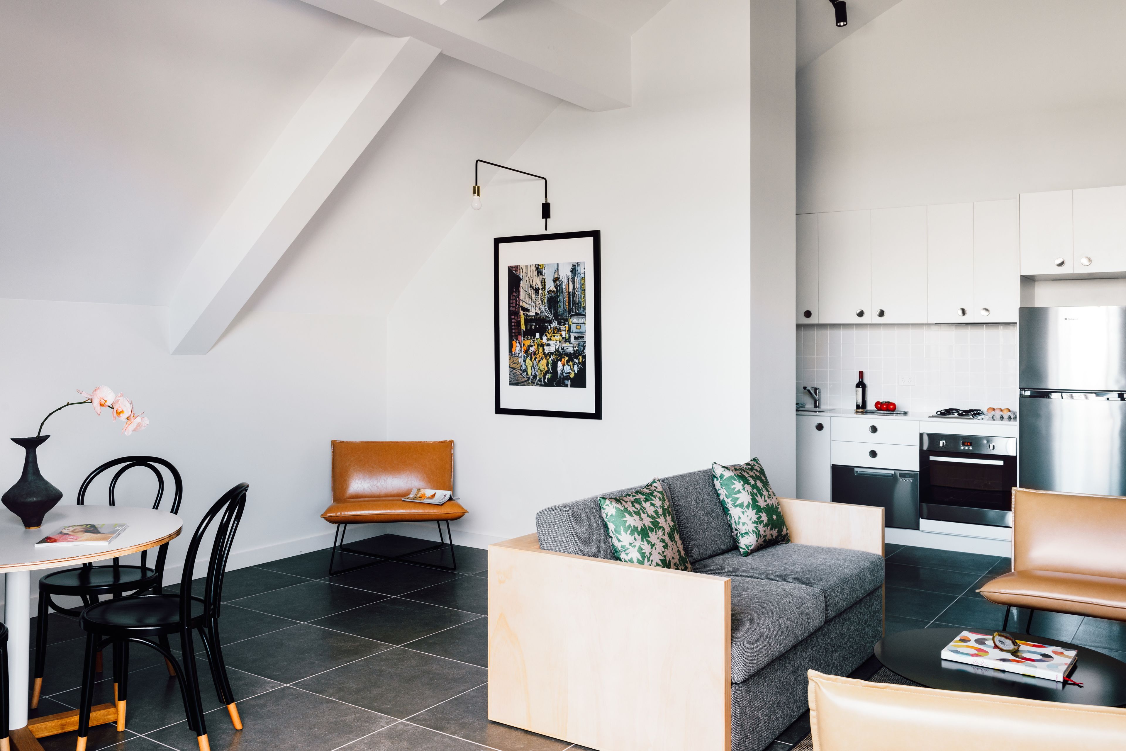 A modern living room with a grey sofa, a coffee table, and a dining area with a white table and black chairs. There is a kitchen in the background with white cabinets and a stainless steel refrigerator.