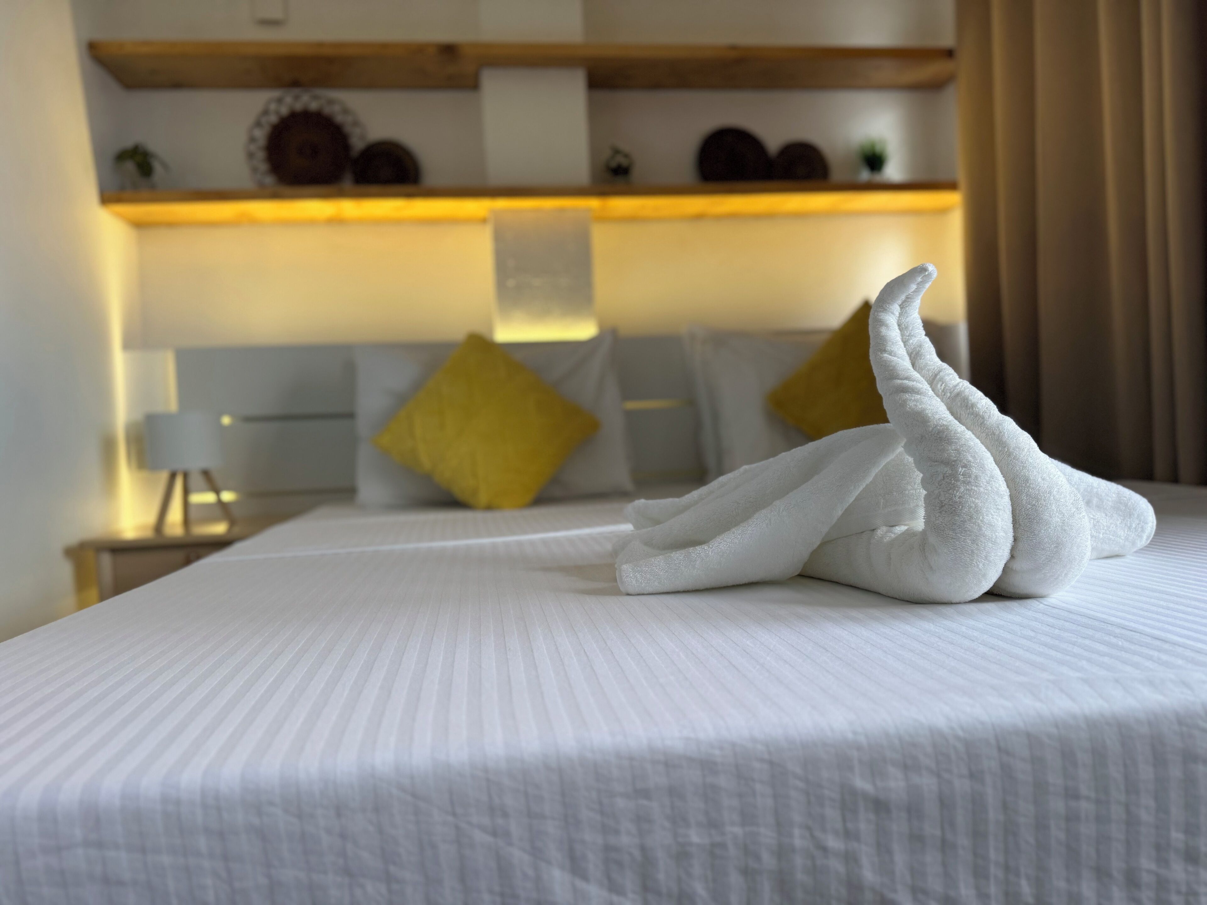 A neatly made bed with a swan towel arrangement, a bedside lamp, and a shelf with decorative items.