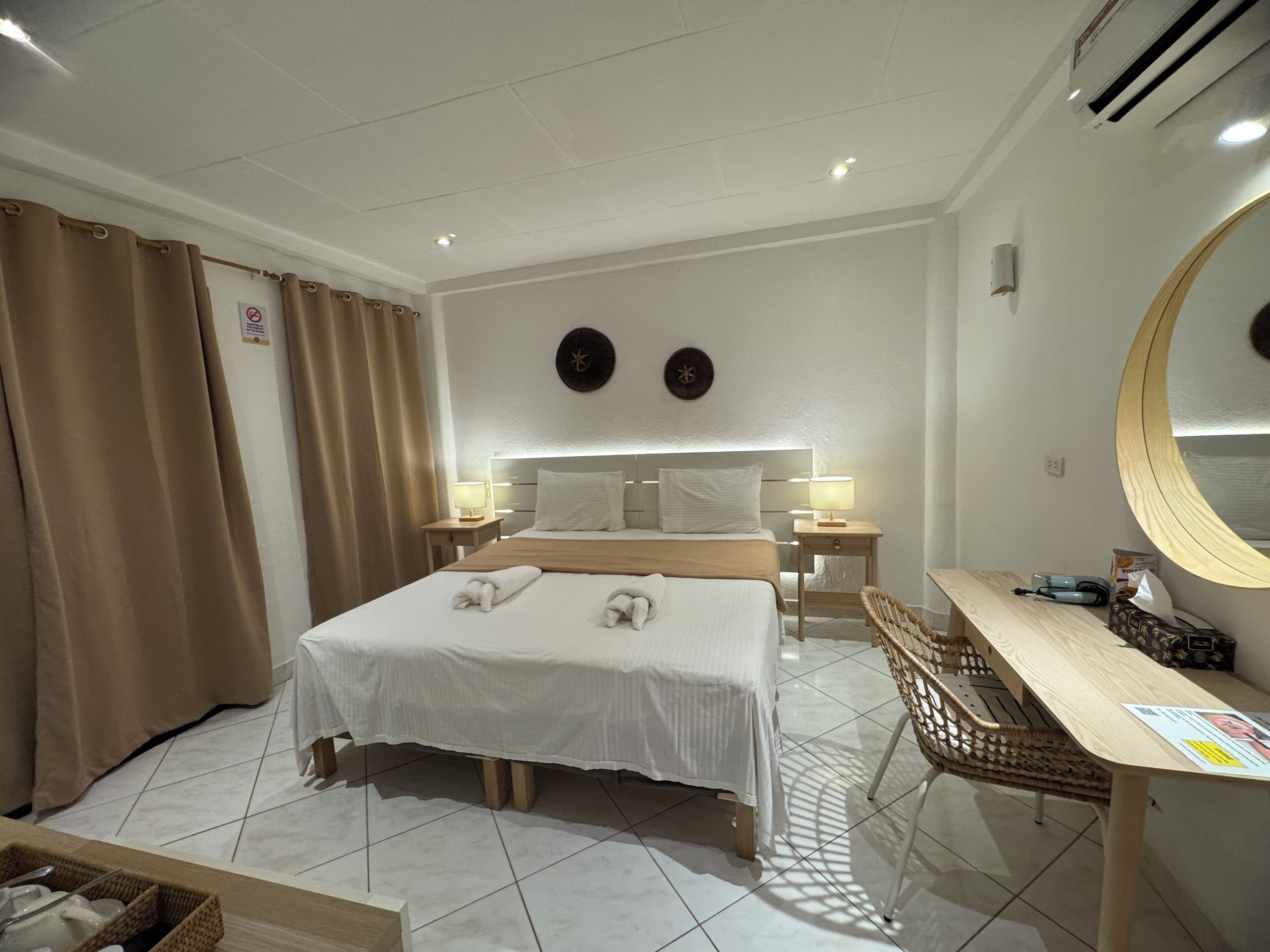 Ein Hotelzimmer mit einem Bett, Nachttischen, einem Schreibtisch, einem Sessel, einem Spiegel und einer Wanduhr.