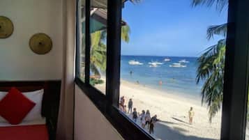 A beach view from a room with a bed and a window overlooking the beach, palm trees, and boats.