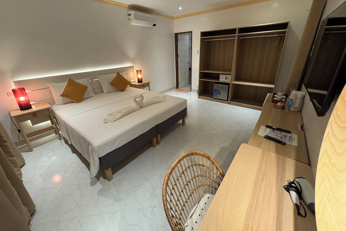 A hotel room with a bed, bedside table, lamp, wardrobe, and a desk with a chair.