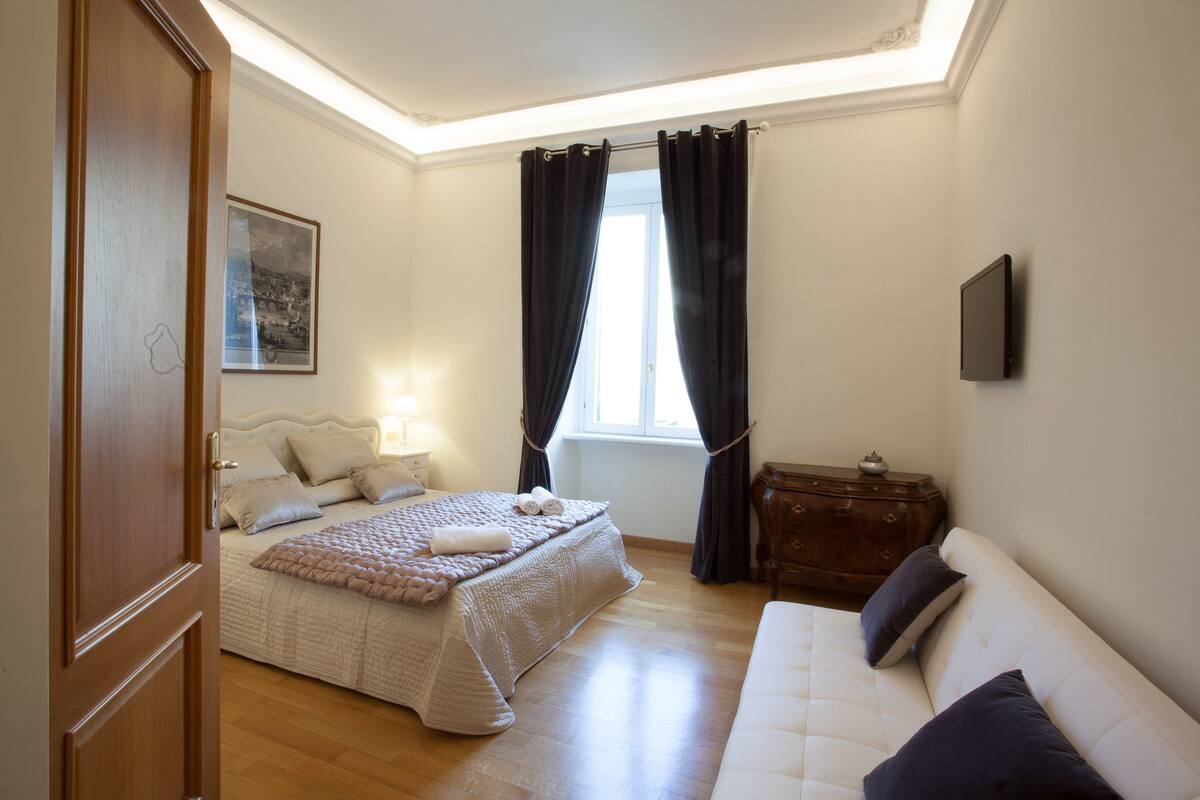 A bedroom with a bed, a sofa, a dresser, a window with curtains, and a wall-mounted TV.