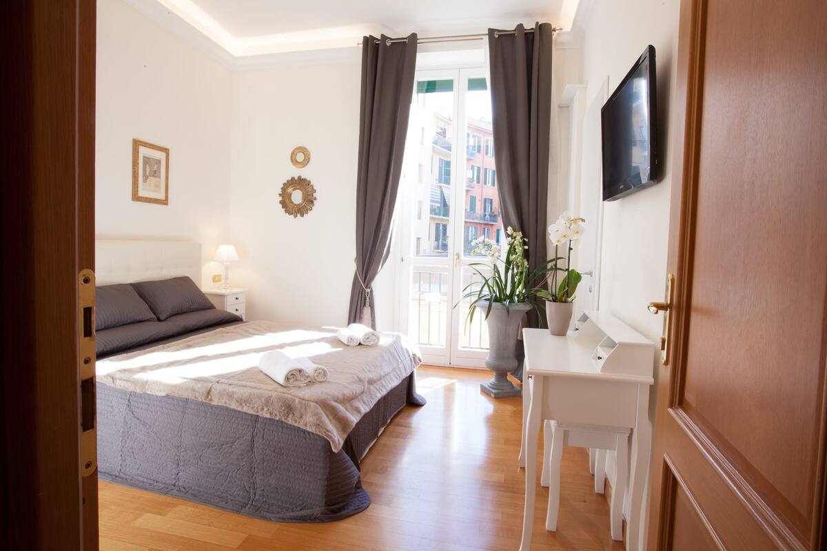 A bedroom with a bed, bedside table, lamp, curtains, a potted plant, a desk, and a TV.