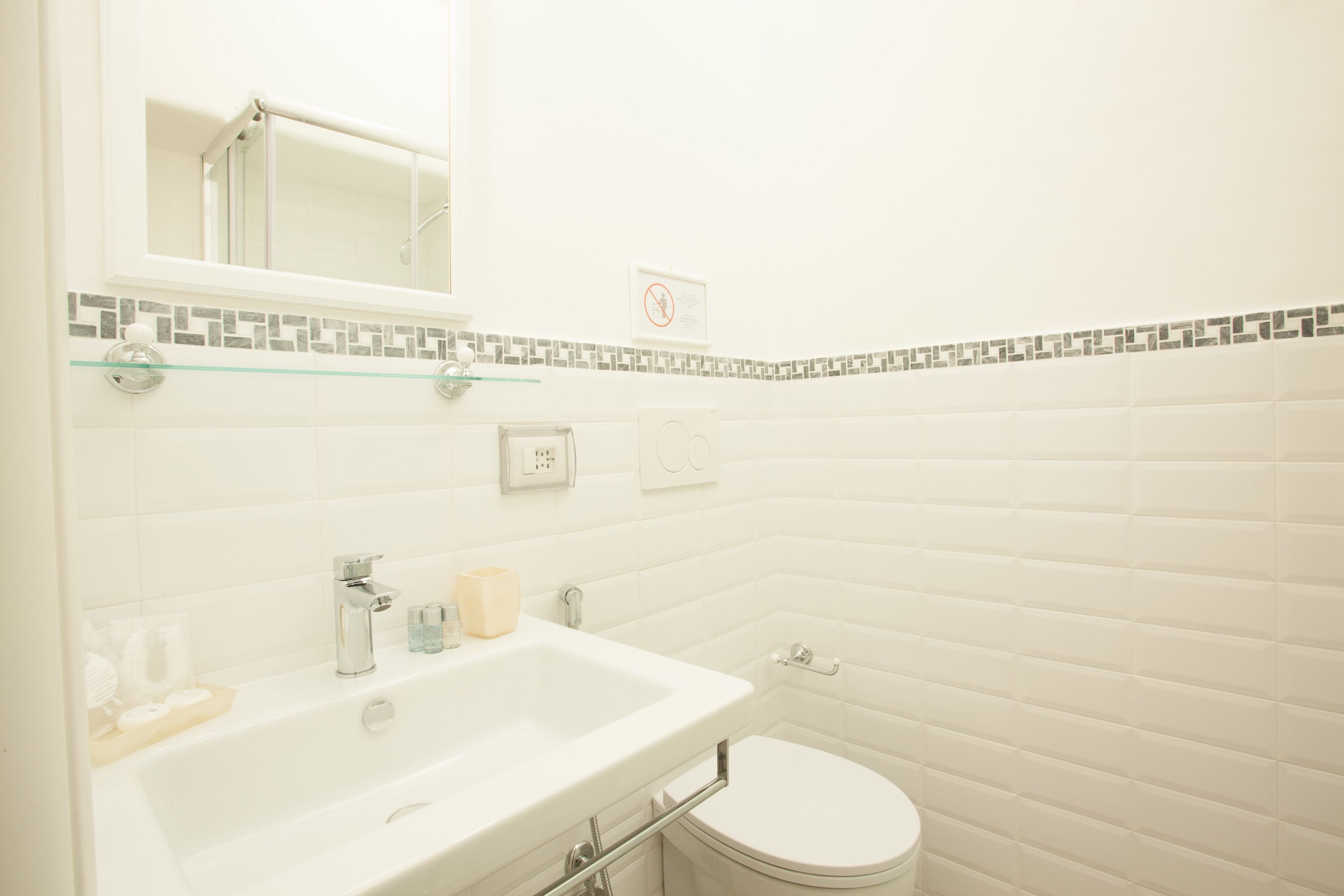 A bathroom with a white sink, mirror, and toilet. There is a glass shelf above the sink and a tiled backsplash.