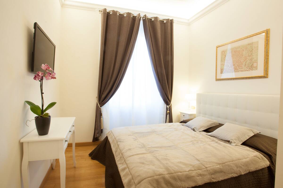 A bedroom with a bed, a bedside table, a potted plant, a wall-mounted TV, and a framed picture.