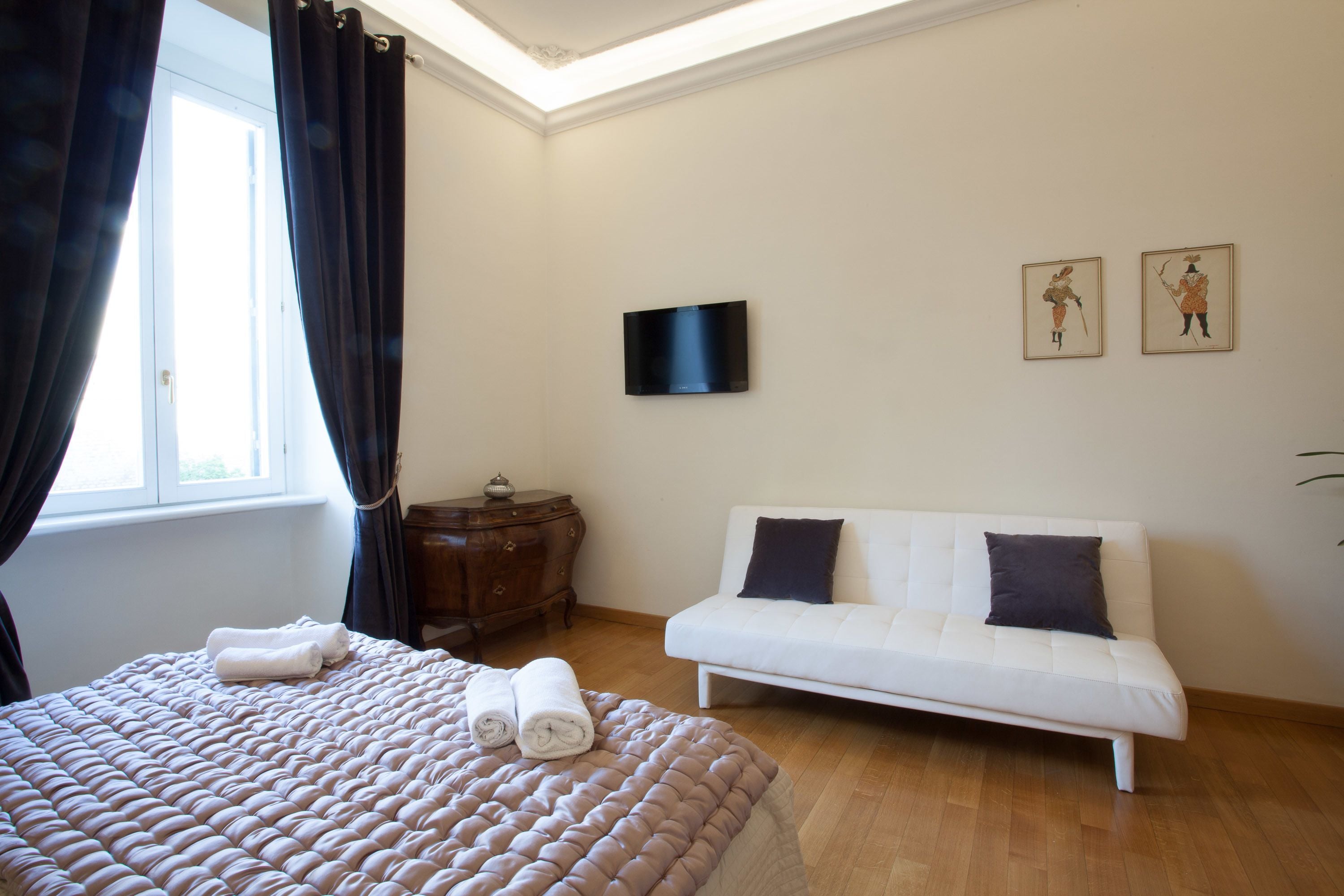 Habitación de hotel con cama, sofá blanco, televisor montado en la pared y cuadros decorativos.