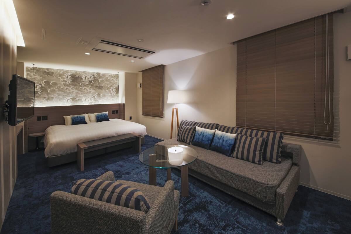 A hotel room with a bed, a sofa, a chair, a glass table, a lamp, and a wall with a textured mural.