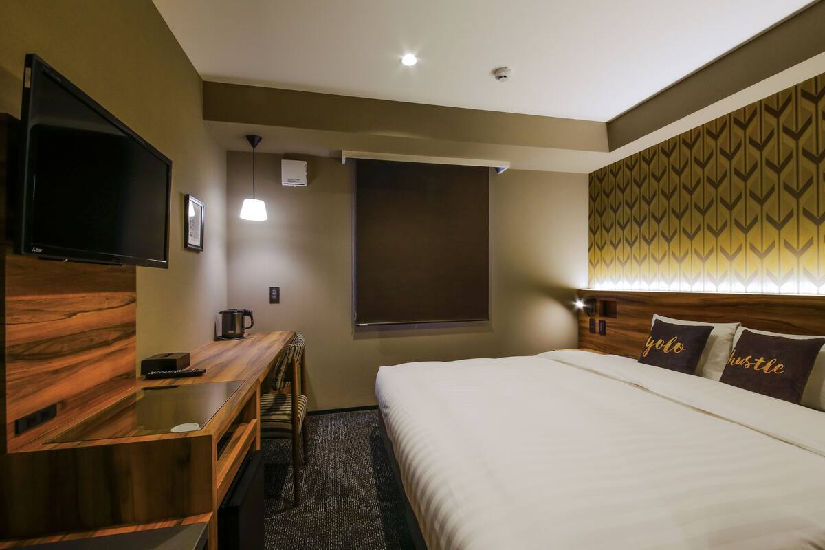 A hotel room with a bed, a TV, a desk with a coffee maker, and a window with a blind.