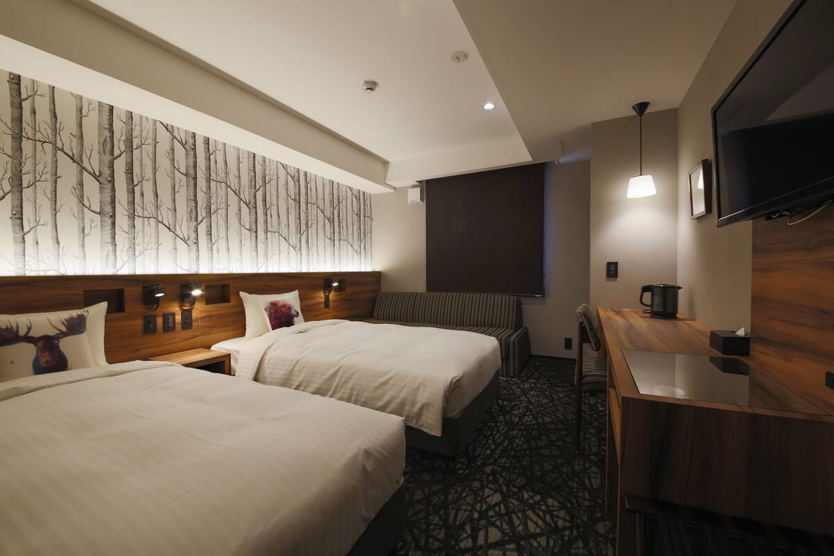 A hotel room with two beds, a wooden headboard, a flat-screen TV, a desk with a coffee pot, and a wall-mounted TV.