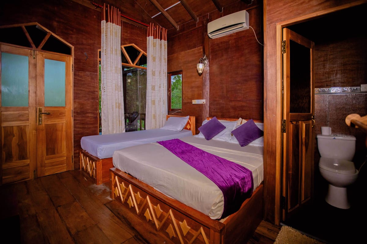 A wooden room with a bed, a window with curtains, an air conditioning unit, and a toilet.