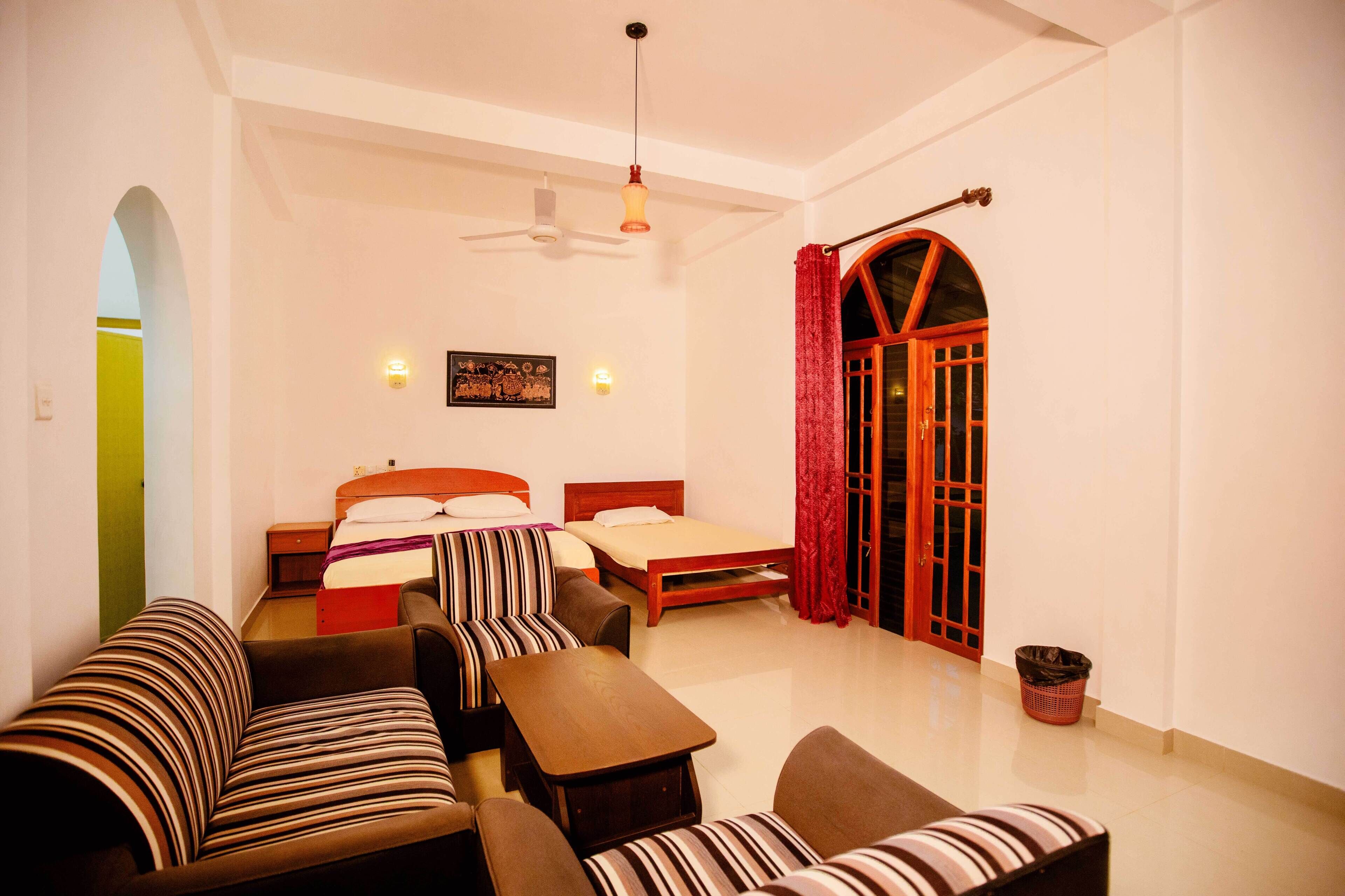 A hotel room with a bed, two sofas, a table, a ceiling fan, and a red curtain.