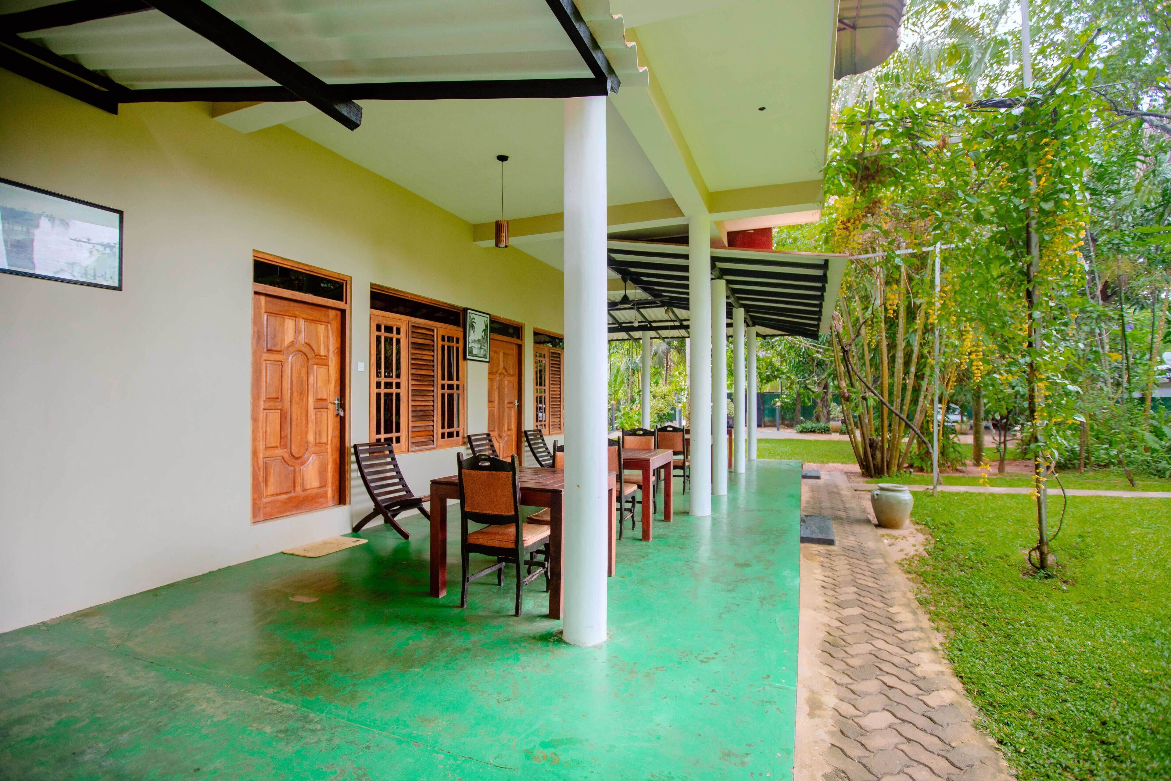 A covered outdoor area with wooden tables and chairs, a paved floor, and a garden with trees and a pathway.