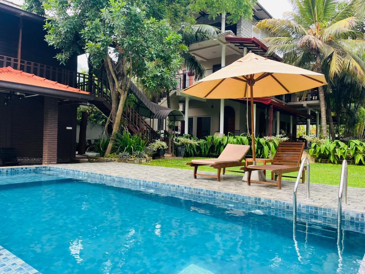 A swimming pool with lounge chairs and an umbrella, surrounded by greenery and a building with a red roof.