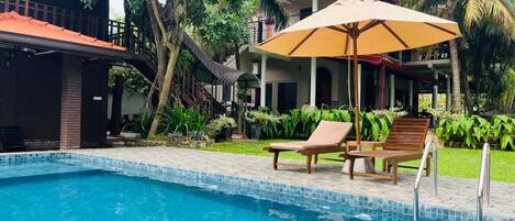 Une piscine entourée de verdure, avec des chaises longues et un parasol, et un bâtiment doté d’un toit rouge.