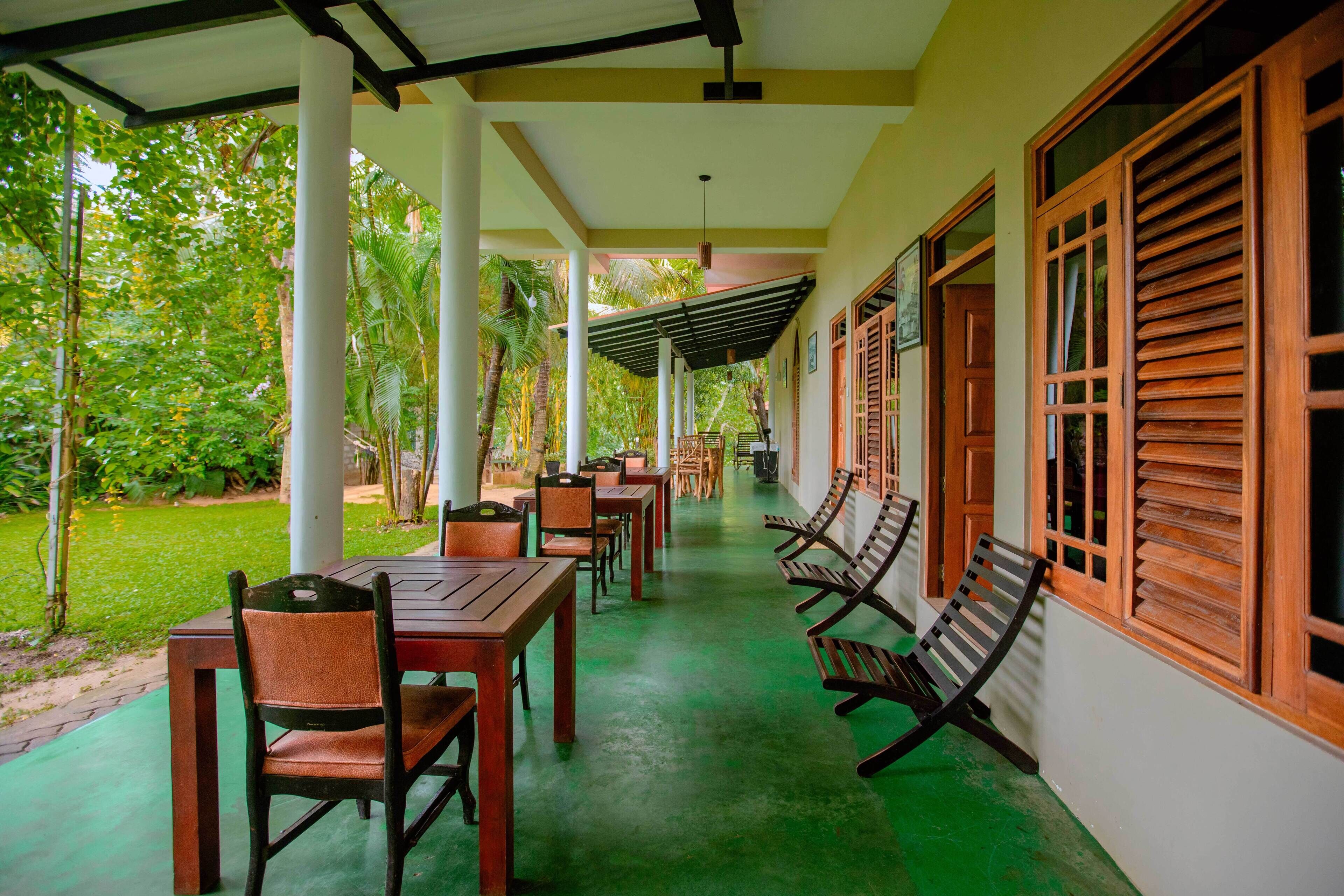 A covered outdoor area with tables and chairs, surrounded by greenery and a building with wooden shutters.